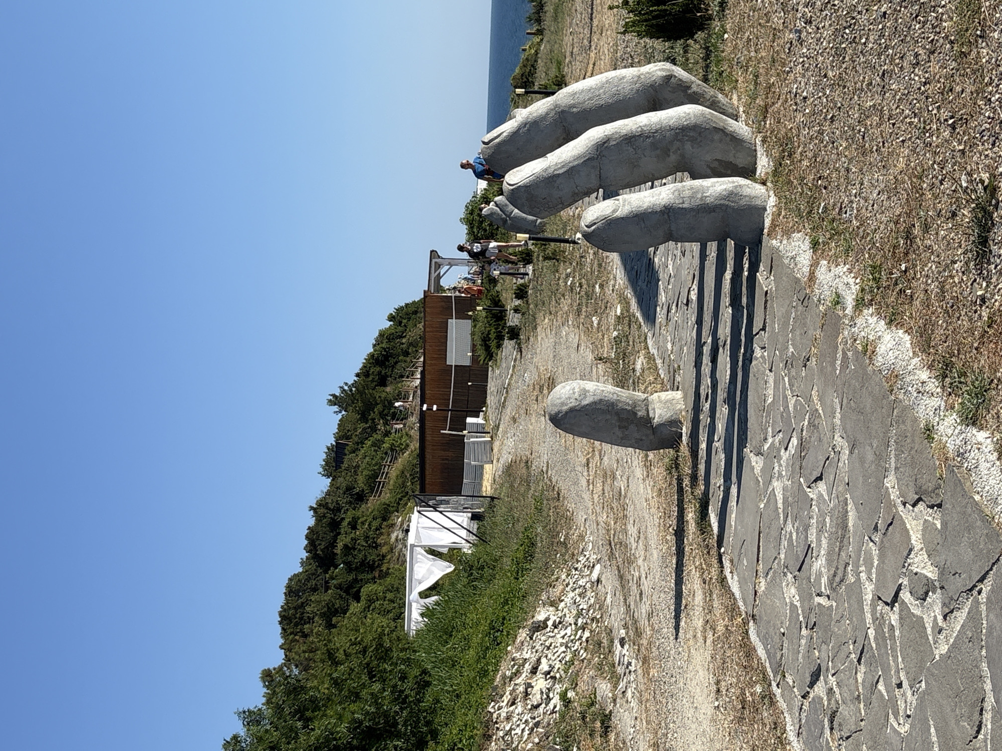The image depicts a large, stone sculpture of a hand, with its fingers extending upwards and casting shadows on a stone pathway. The sculpture appears to be made of concrete or a similar material and is situated outdoors. Several people are visible interacting with the sculpture, with some standing on or near the fingers, and others walking along the path. In the background, there is a building with a wooden exterior, possibly a restaurant or cafe, with white tents or awnings set up nearby. Lush green trees and vegetation cover the hillside behind the building, and the blue expanse of the sea can be seen in the distance. The sky is clear and bright blue, suggesting a sunny day. The overall scene conveys a sense of a tourist attraction or a public space designed for leisure and enjoyment. The time of day appears to be daytime, likely late morning or early afternoon, given the angle of the shadows. The location is identified as Kabardinka, Russia. No visible text is present in the image.