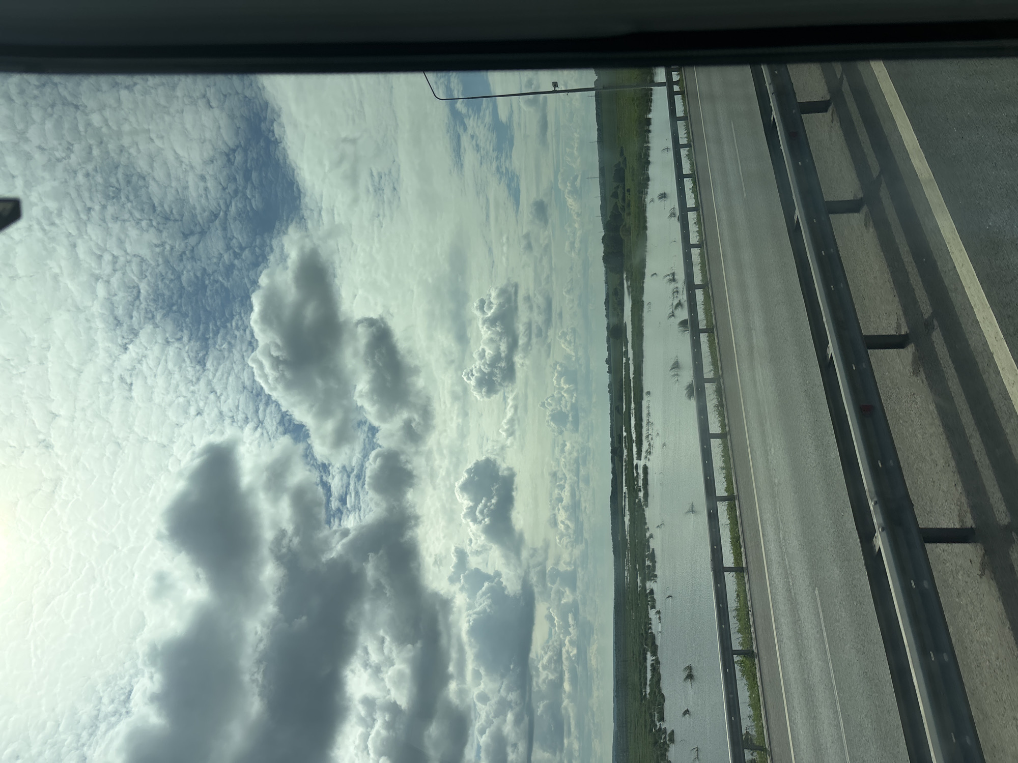 The image depicts a highway scene with a wide, calm body of water adjacent to the road, likely a river or a lake. The water is dotted with green vegetation, appearing to be partially submerged. The sky is filled with dramatic clouds, a mix of dark, heavy storm clouds and lighter, wispy formations, suggesting dynamic weather. A metal guardrail runs along the edge of the highway, separating it from the water. A streetlamp is visible on the right side of the frame, near the horizon. The overall impression is one of a natural landscape viewed from a moving vehicle. Given the provided context, this scene is located in the city of Menzelinsk, Russia.