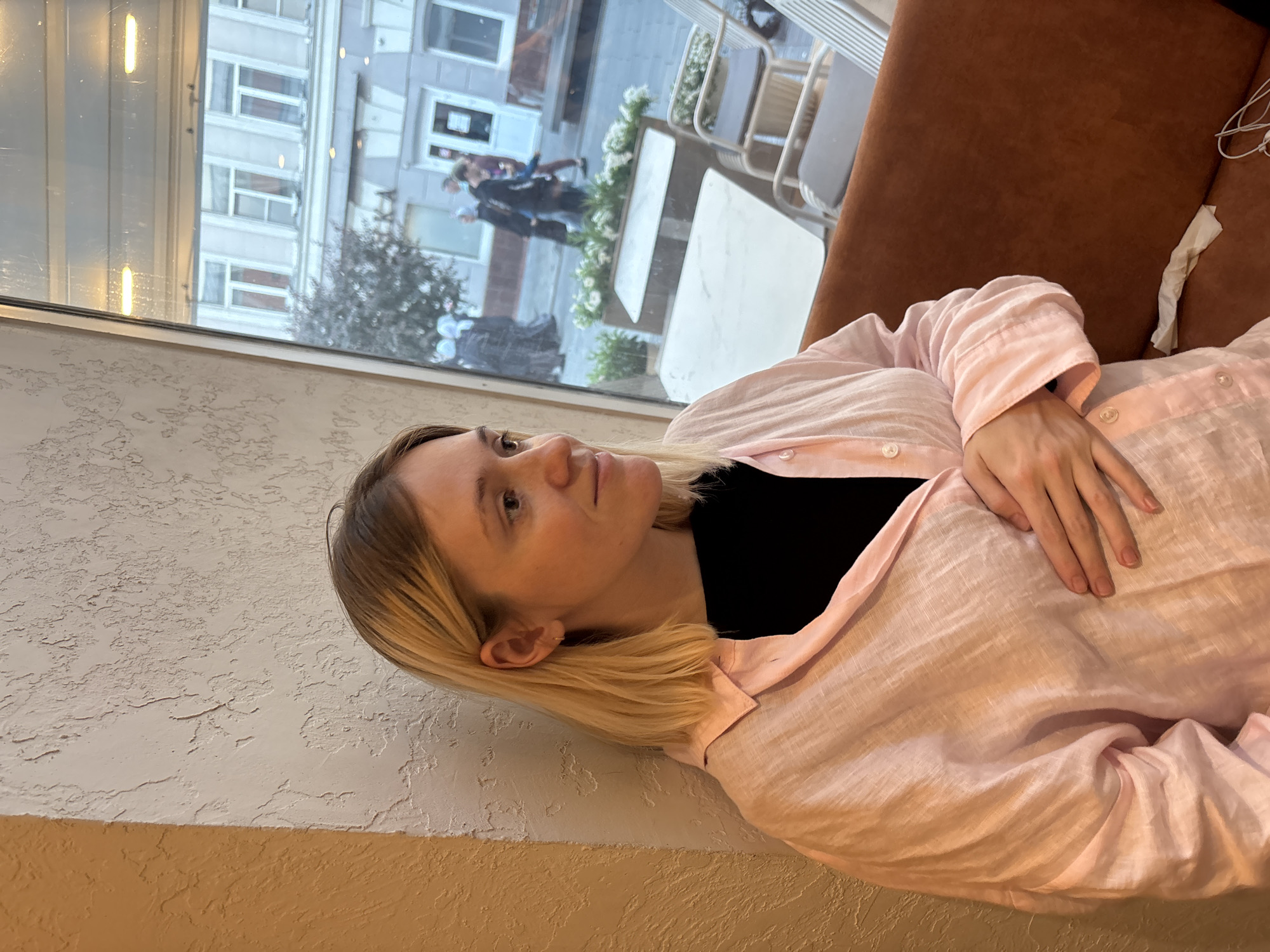 A young woman with blonde hair is sitting by a window. She is wearing a pink open shirt over a black top and has her hand on her stomach. Outside the window, people are walking on a street with buildings. The location appears to be a cafe in Nizhny Novgorod, Russia, given the architecture visible through the window. The woman is looking away from the camera, towards the window.