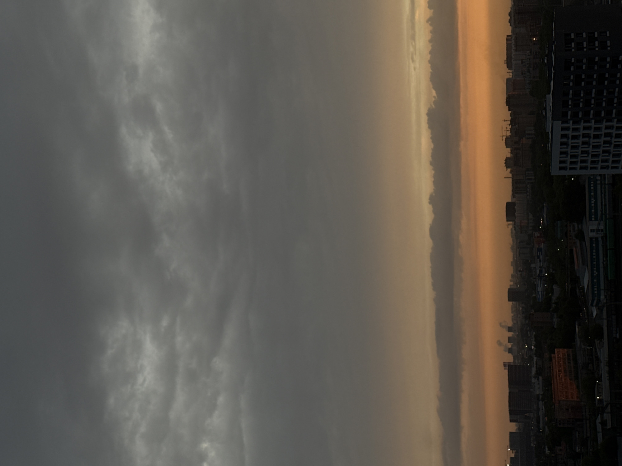 The image presents an aerial view of what appears to be the city of Lyubertsy, Russia, though the precise location within the city is indeterminable. The scene is dominated by a sky that is mostly overcast with dark, grey clouds, indicating possible inclement weather. To the right of the image is a skyline of buildings, likely residential and commercial, some appearing modern with high-rise structures. There is an orange glow separating the city from the sky, possibly indicative of the setting sun obscured by the clouds, and there are city lights that provide illumination as well. There are no identifiable people or specific actions visible. The image conveys an urban environment during the transition from day to night, with a focus on atmospheric conditions and city landscape.