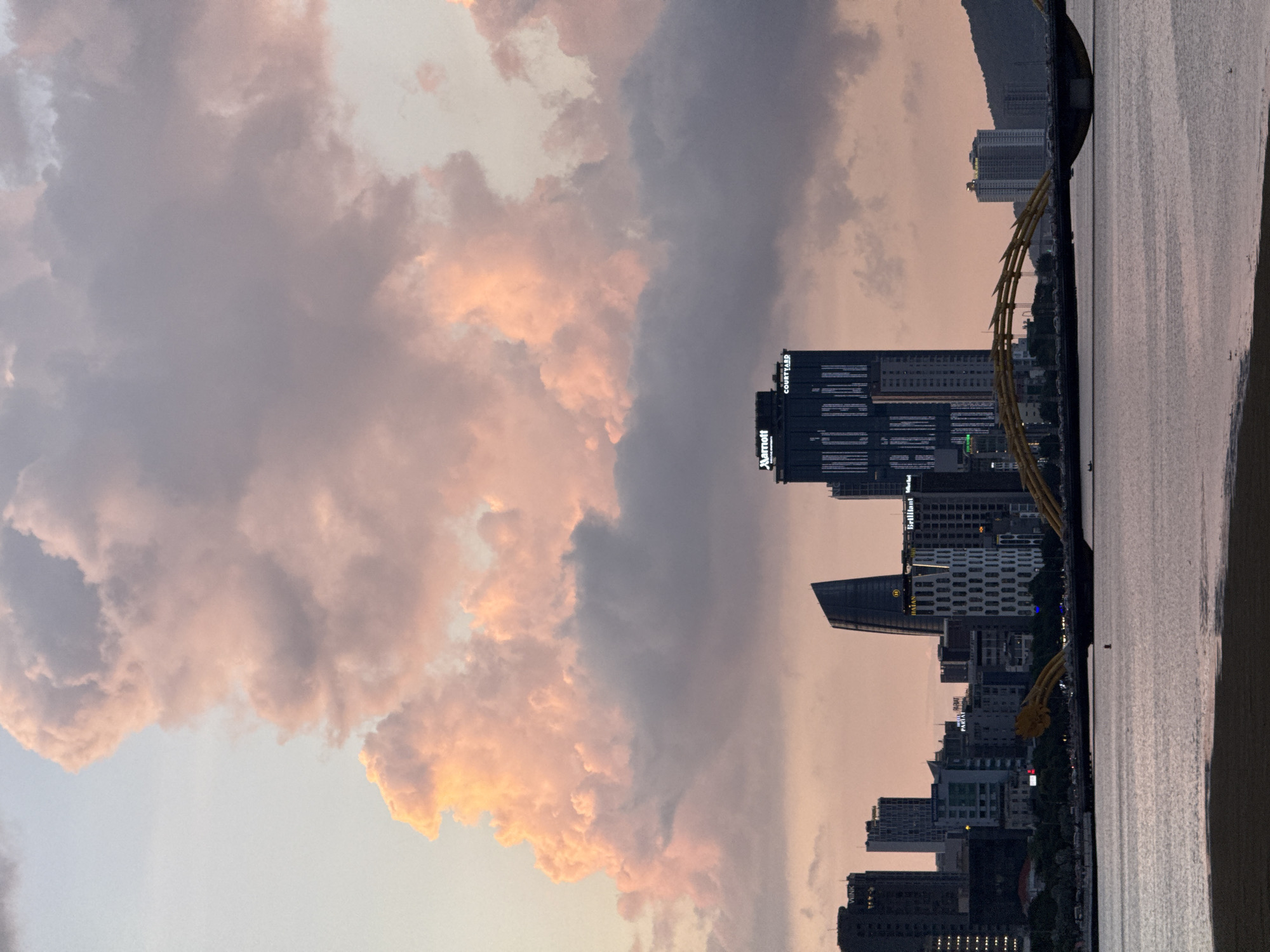 This image captures the Dragon Bridge in Da Nang, Vietnam, at sunset. The sky is filled with large, dramatic clouds, illuminated in soft pinks and oranges by the setting sun. In the foreground, a body of water reflects the sky and the cityscape. The Dragon Bridge, a prominent landmark, spans the river with its distinctive dragon-shaped arches. The city skyline features a collection of modern buildings, including the Marriott and Courtyard by Marriott hotels, with their signs visible. The overall scene is serene, with the warm hues of the sunset creating a picturesque view of the city.