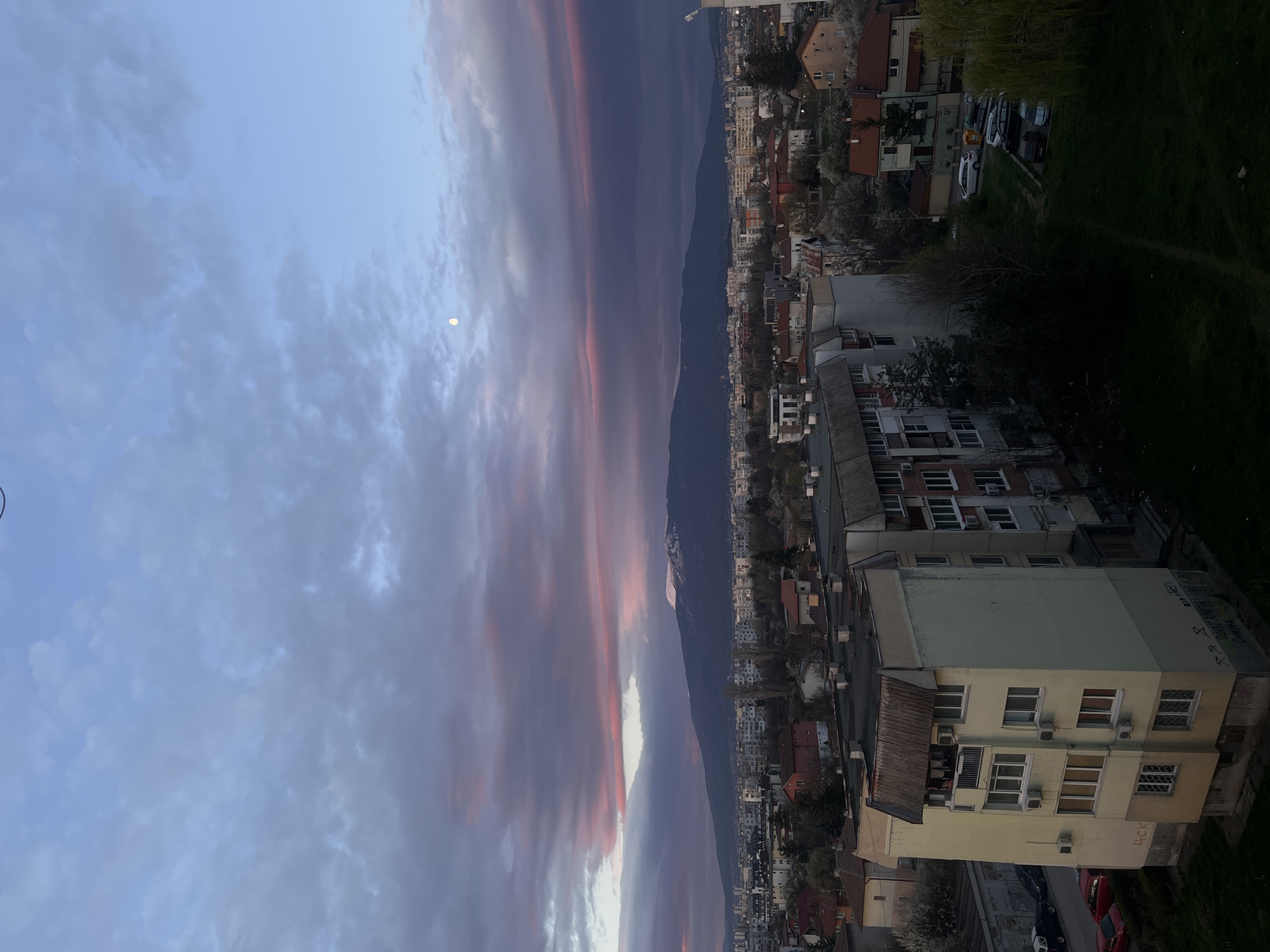 The image shows a cityscape with mountains in the background and a colorful sunset sky. In the sky, there is a visible moon. The city consists of buildings of various heights and styles, with some trees interspersed among them. Some houses are visible, with a red roof. A tall, dark mountain range runs between the sunset and the city. There is a building in the lower right, with several air conditioning units attached. It is difficult to determine if there are people or activities within the frame. The environment appears to be a residential or mixed-use urban area. The location is possibly Sofia, Bulgaria.