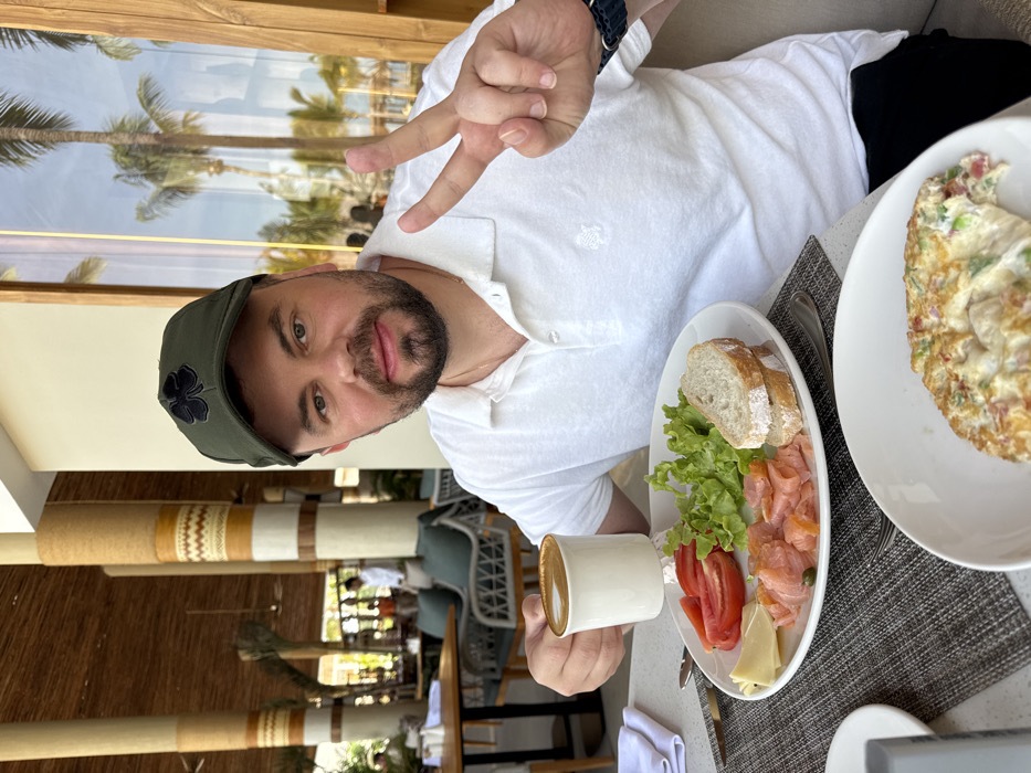 A man in a green hat and white polo shirt sits at a table. He holds up two fingers in a peace sign and a cup of coffee. Two plates with food are visible: one with bread, lettuce, salmon, tomatoes, and cheese, and the other with an omelet. Palm trees are visible in the background through a window.