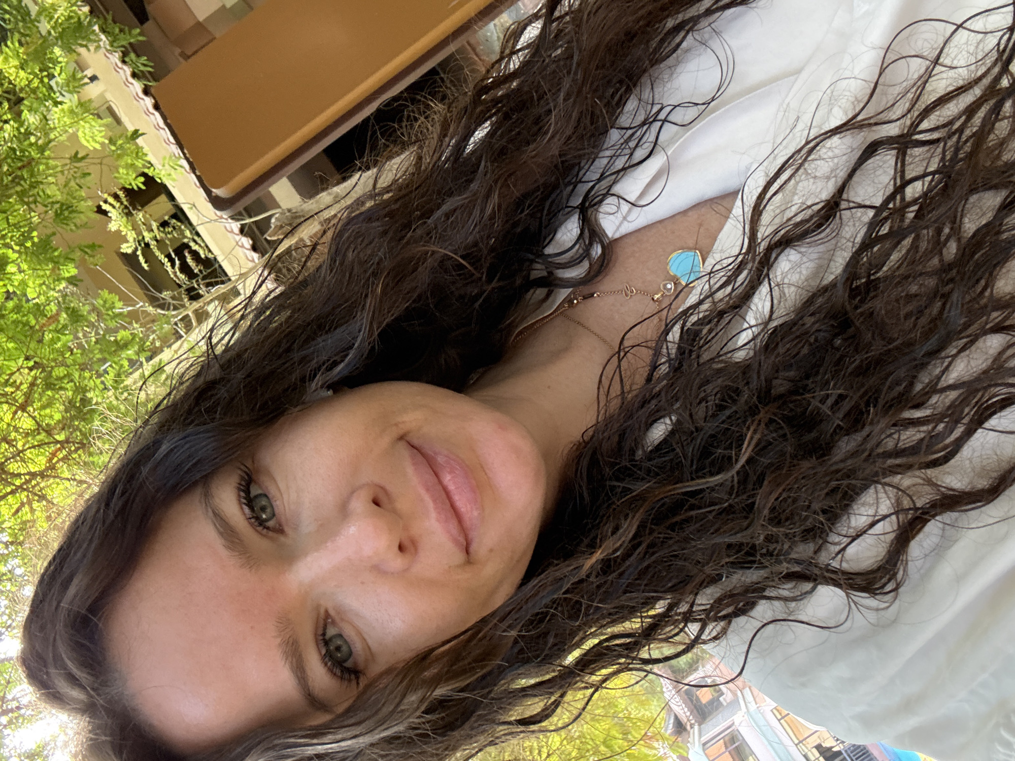 A selfie of a woman with dark, wavy hair and green eyes is taken outdoors. She is wearing a white shirt and a necklace with a blue heart-shaped pendant. The background shows lush green trees, a building with balconies, and a tan-colored wall or structure. The image is well-lit, suggesting it was taken during the day. The woman is smiling, and her hair appears wet, possibly indicating she has recently been swimming or showering. The setting suggests a tropical or resort-like environment. The specific location in Dubai, United Arab Emirates, cannot be determined from the visual information present.