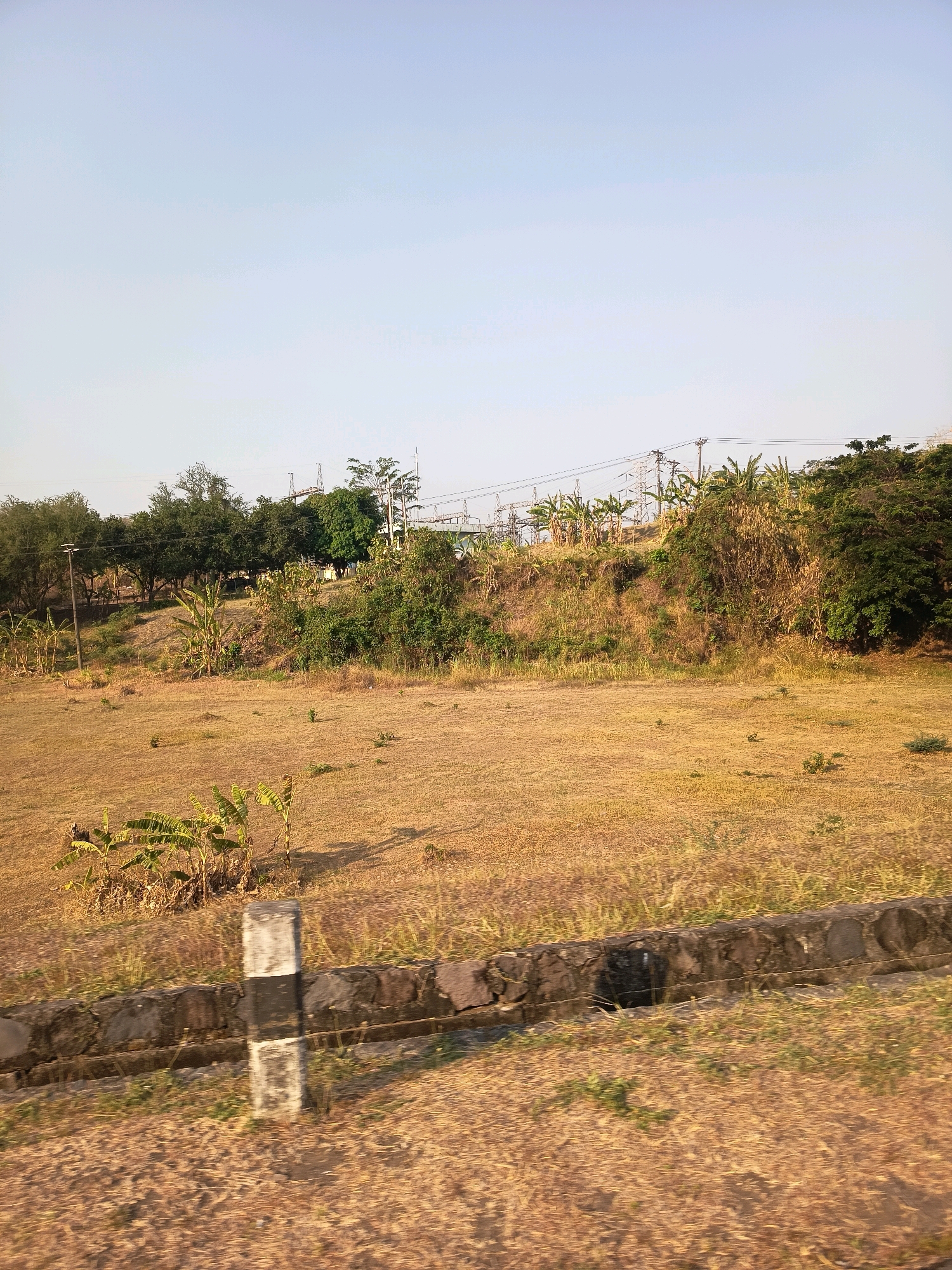 The image captures an outdoor scene in the city of Kalangbancar, Indonesia. In the foreground, a dry, grassy field stretches out, bordered by a low stone wall. A black and white post stands prominently near the wall. Beyond the field, a gentle slope rises, covered in dry grass and scattered vegetation. Towards the top of the slope and in the background, a dense line of trees and foliage is visible. Electrical power lines and transmission towers are situated on the elevated terrain, suggesting the presence of infrastructure. The sky above is a clear, pale blue, indicating fair weather conditions. The overall impression is one of a rural or semi-rural landscape on a sunny day.