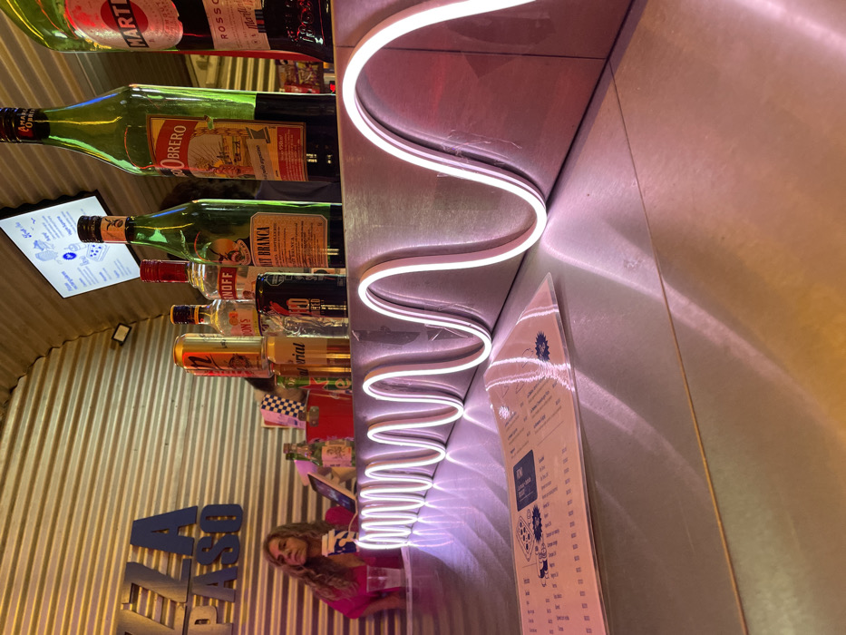 A bar shelf displays various liquor bottles, including Martini Rosso and Obrero Argentino. A white neon light snakes along the shelf's edge, casting a bright glow. A clear menu stands nearby, showcasing text and illustrations. A woman in a pink top and blue and white shorts is visible at the bottom. The wall behind the shelf is corrugated metal, while the wall next to it is light pink.