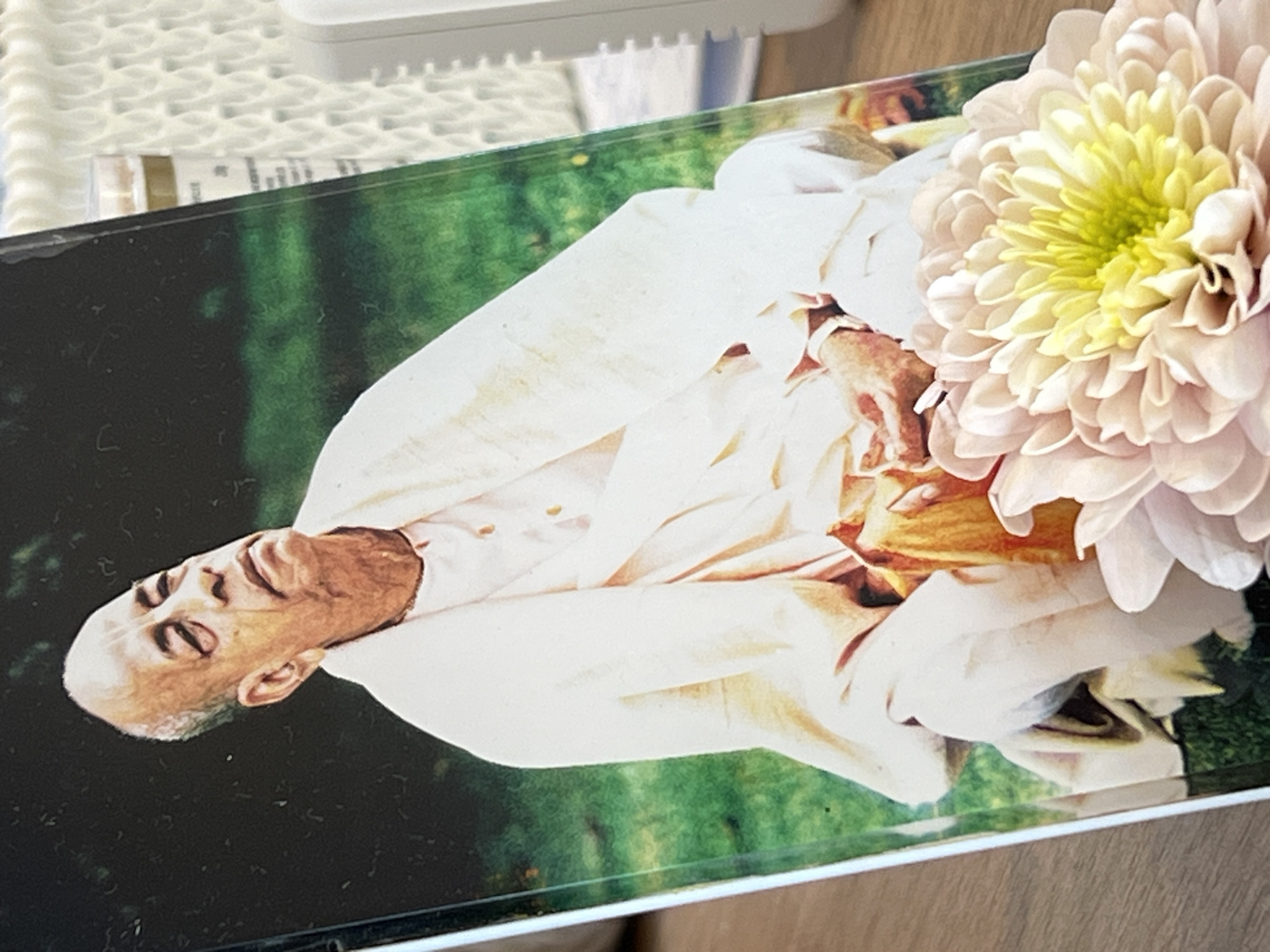 The image shows a close-up of a book with a portrait of an elderly man on the cover, dressed in light-colored robes. He is seated, with his head tilted upwards. In the foreground, a pale pink and yellow chrysanthemum is partially obscuring the bottom of the book. The background is out of focus, showing green foliage and a glimpse of a white woven texture and a white electronic device. The book is positioned on a wooden surface. There is no information within the image to suggest any specific location in Bunbeg, Ireland.