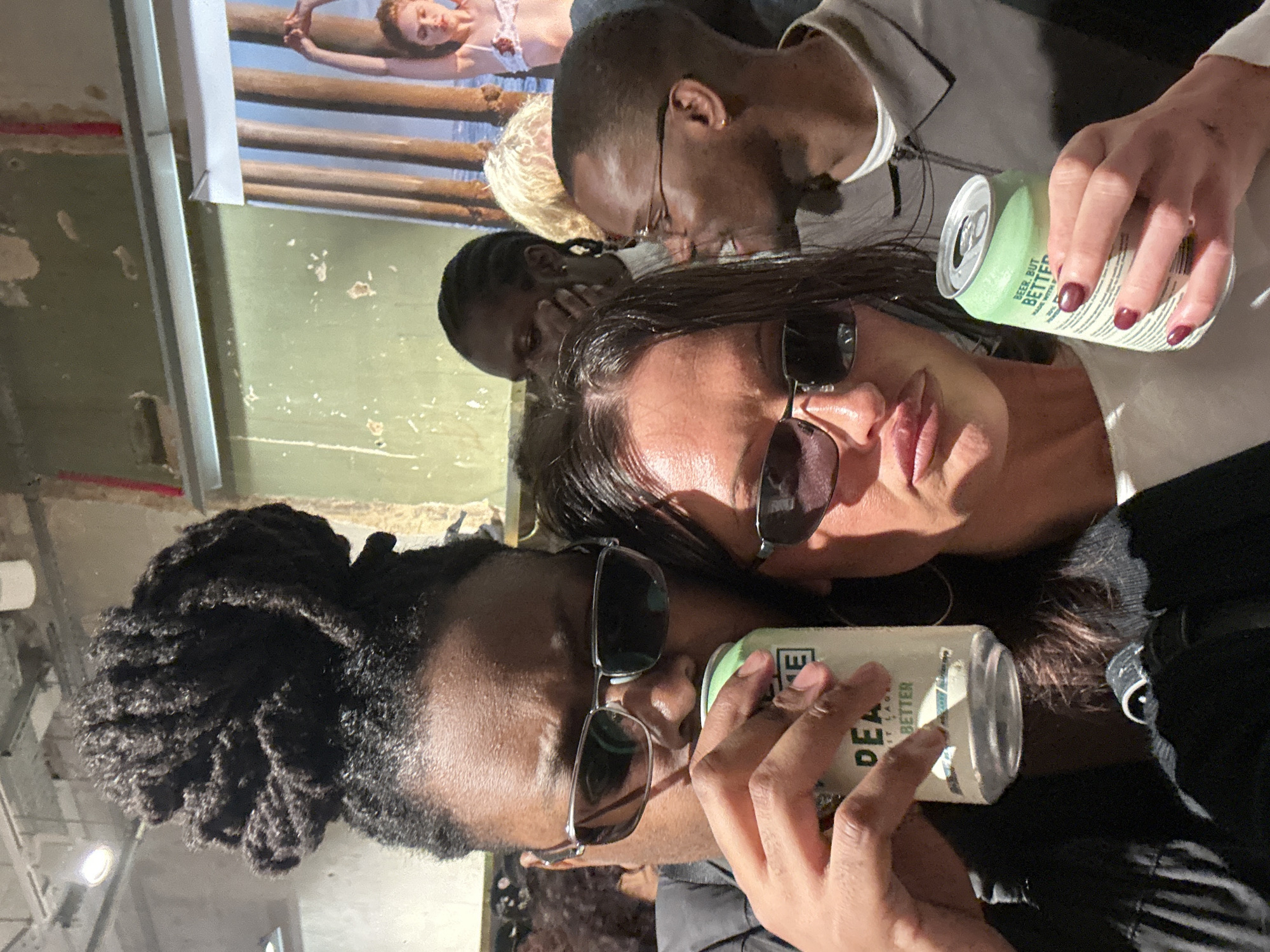 The image, oriented correctly, features four individuals in an indoor setting, possibly a bar or event space in London, United Kingdom. In the foreground, a person with dreadlocks and dark sunglasses is seen drinking from a light green can. Beside them, another person with long dark hair and dark sunglasses looks towards the viewer with a slight pout, also holding a light green can in their right hand. Their left hand rests near the first person's shoulder.
Behind these two, a person with short dark hair and glasses is visible, looking downwards. Further in the background, another person is partially obscured. The two visible cans clearly show the text "BEER. BUT BETTER.", with "PEA" and "BETTER" also partially visible on the other can, indicating the same product.
The background features distressed, possibly industrial-style walls with peeling light green/grey paint and hints of red. A vertical white structure is on the left. On the wall in the upper left, a framed picture or poster depicts a person, likely a woman, lying on wooden planks over water. The overall atmosphere appears casual and social, with the subjects appearing relaxed. The lighting suggests an indoor setting with artificial light.