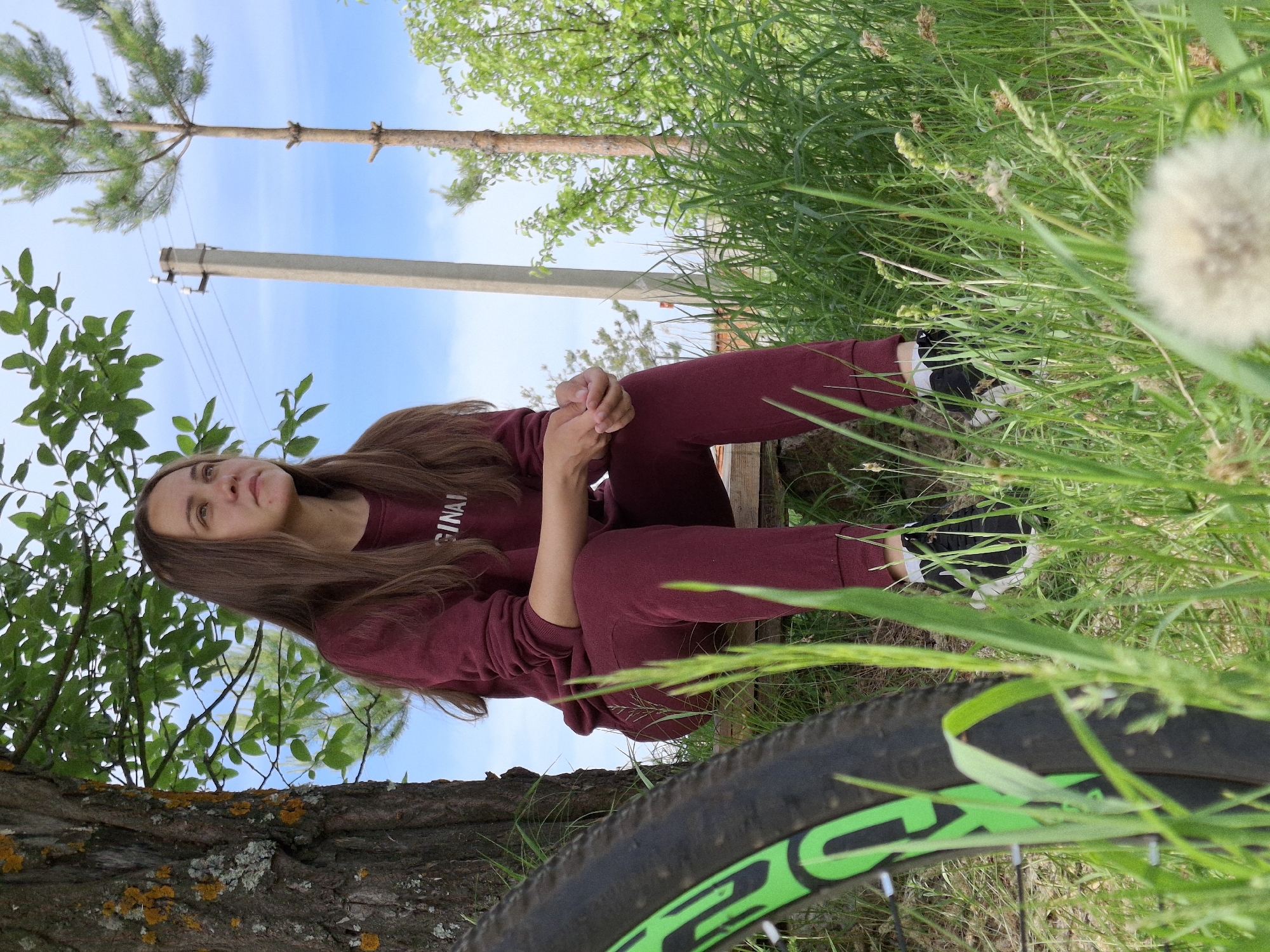 A young woman is sitting on a wooden bench outdoors in a natural setting. She is wearing a maroon colored tracksuit. She sits near a tree in Chaikovsky, Russia. A bicycle tire is prominent in the foreground. Tall grass and a dandelion are also in the foreground. A power pole and trees are in the background under a blue sky.