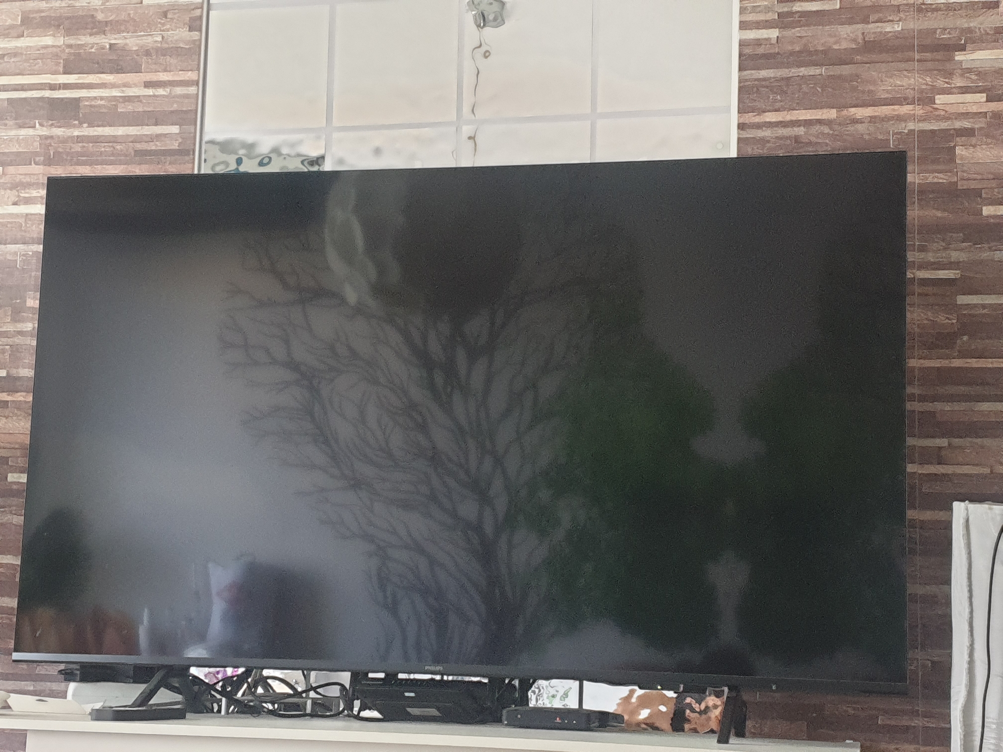 An electronic television screen is positioned centrally on a white shelf in front of a textured brick wall. The screen displays a reflection of a leafless tree and some blurry green shapes, suggesting it is off or displaying a subdued image. Below the screen, a black electronic device and a cluster of black cables are visible. To the right of the television, a white towel or fabric hangs vertically. The overall environment appears to be an indoor living space. There is no indication of the location being Sofia, Bulgaria.