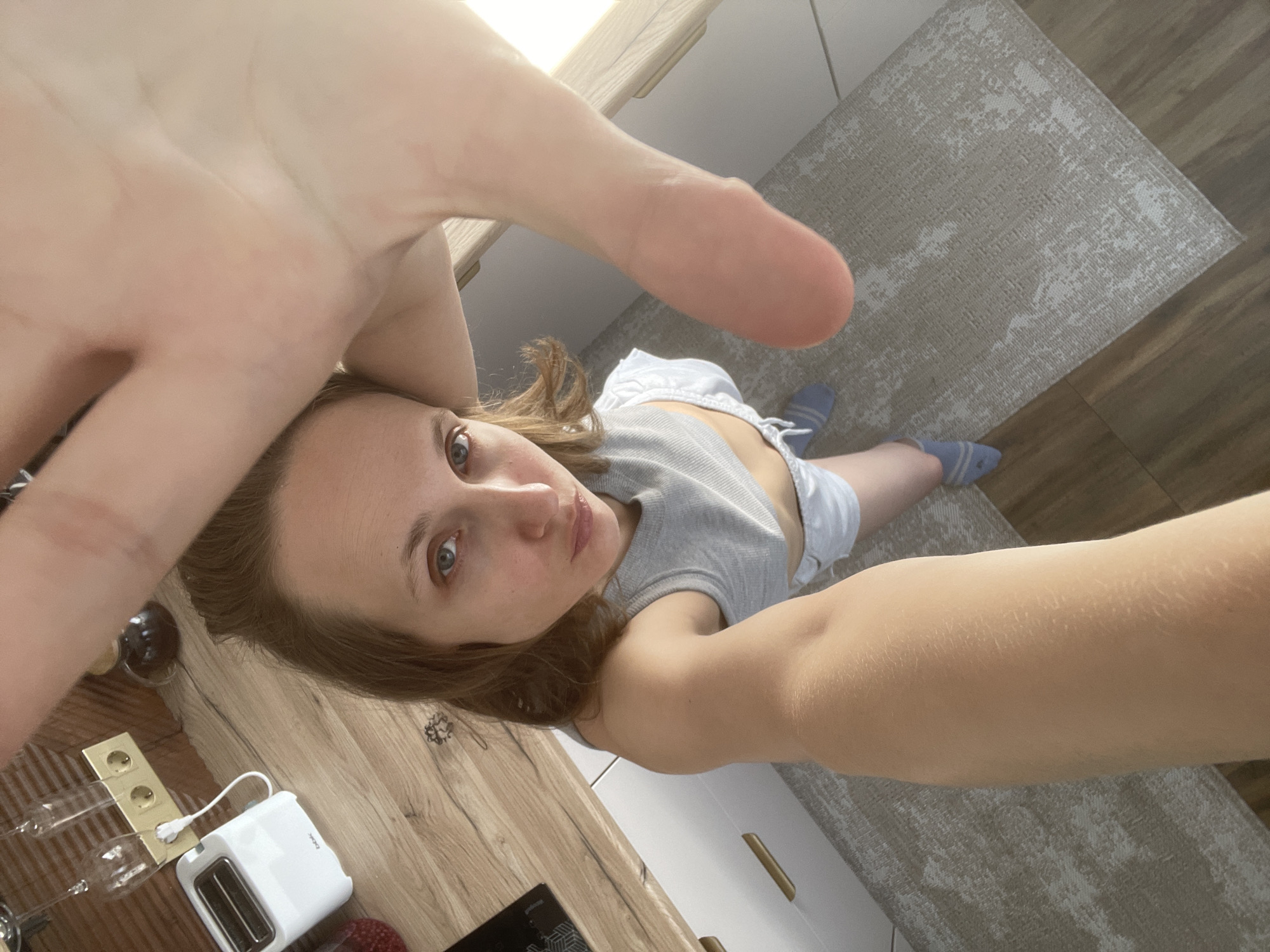 A woman is taking a selfie in a kitchen. The woman has light skin, brown hair, and blue eyes. She is wearing a gray tank top and white shorts. Her right arm is extended toward the camera. A white toaster is on the counter to her left. On the floor is a rug, and wood flooring is around the rug. The location is Unknown, Unknown.