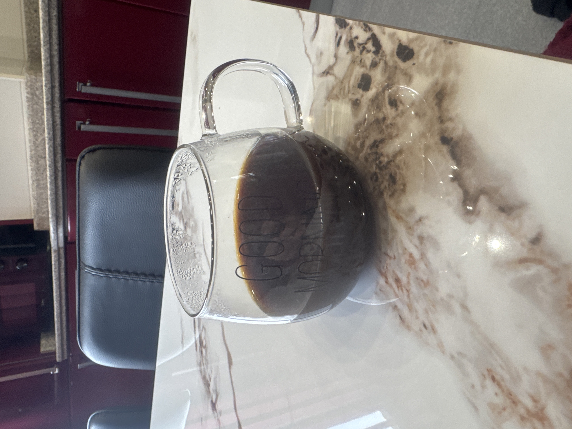 The image displays an indoor scene, likely a kitchen or dining area in Tbilisi, Georgia. A clear glass mug, half-filled with a dark brown liquid (consistent with coffee), is centrally positioned on a white and brown marble-patterned tabletop. The mug has the words "GOOD MORNING" printed in dark letters on its side, visible through the liquid. Condensation is present on the interior rim of the mug. Behind the table, a dark grey or black upholstered chair is partially visible. Further back, a section of a kitchen with dark red cabinets and a light speckled countertop is discernible. Light reflections are prominent on the table surface and the glass mug. No individuals are visible.