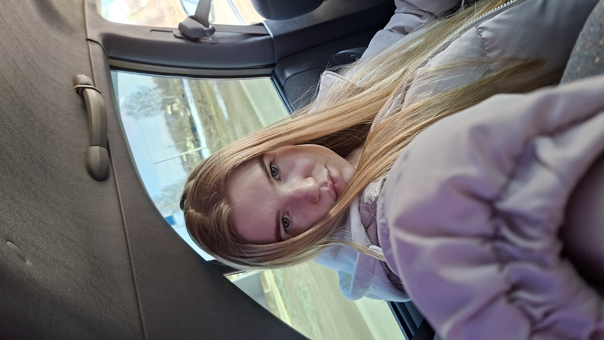 A young woman with long, strawberry blonde hair and fair skin sits in the back of a car. She is wearing a light-colored, puffy jacket. The car's interior is visible, including a grab handle on the interior side panel. Outside the window, a blurred landscape can be seen, suggesting movement. The location is Гаревая, Russia.