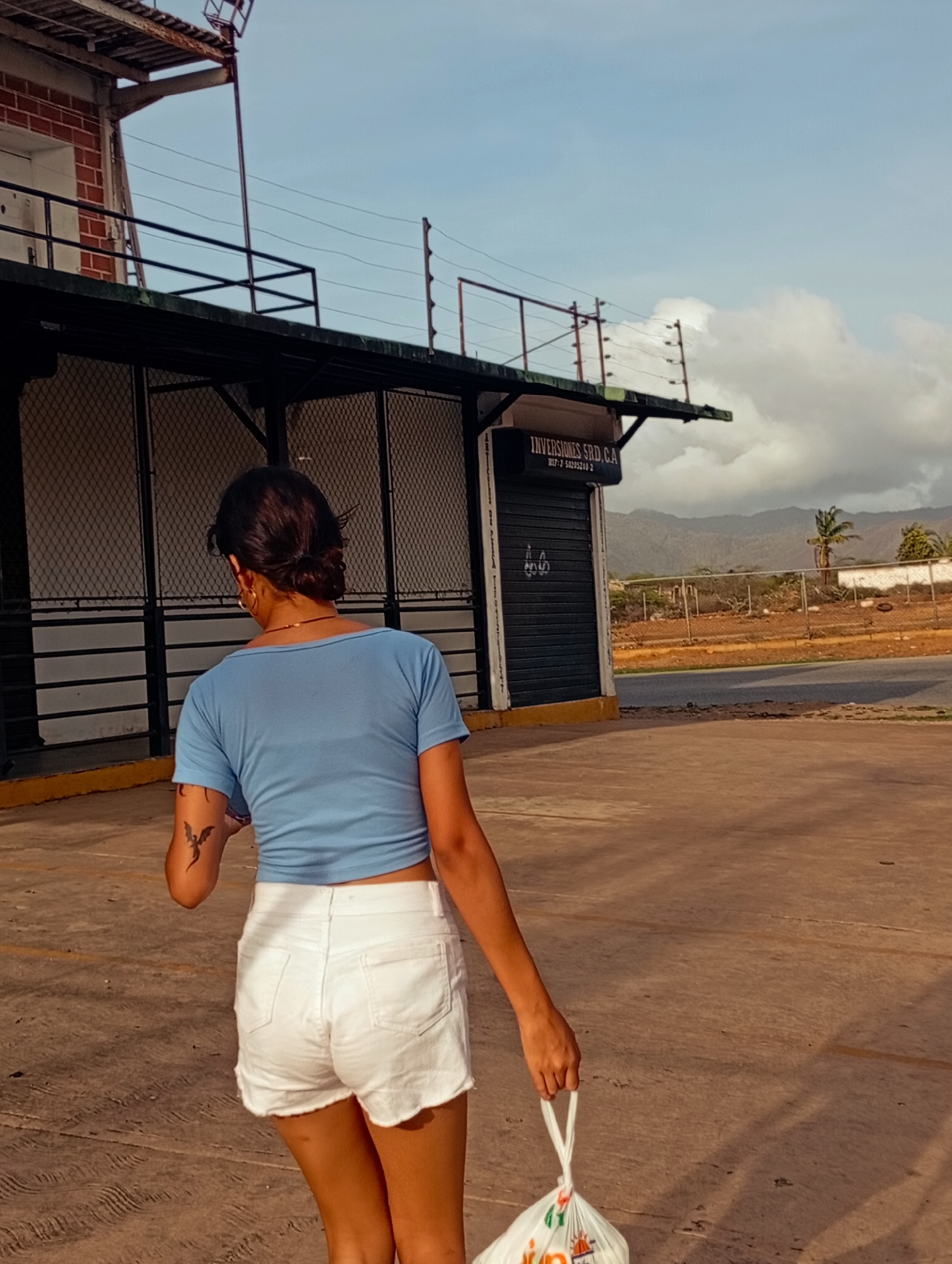 A person is walking away from the camera, carrying a white plastic bag in their right hand. They are wearing a light blue crop top and white denim shorts. A tattoo of a dragon is visible on their left forearm. The person is on a paved outdoor surface, likely a street or walkway, in front of a building with a dark corrugated metal shutter and a sign that reads "INVERSIONES SRD, CA". The background features a chain-link fence, dry vegetation, a palm tree, and mountains under a cloudy sky. The scene appears to be in Juan Griego, Venezuela.