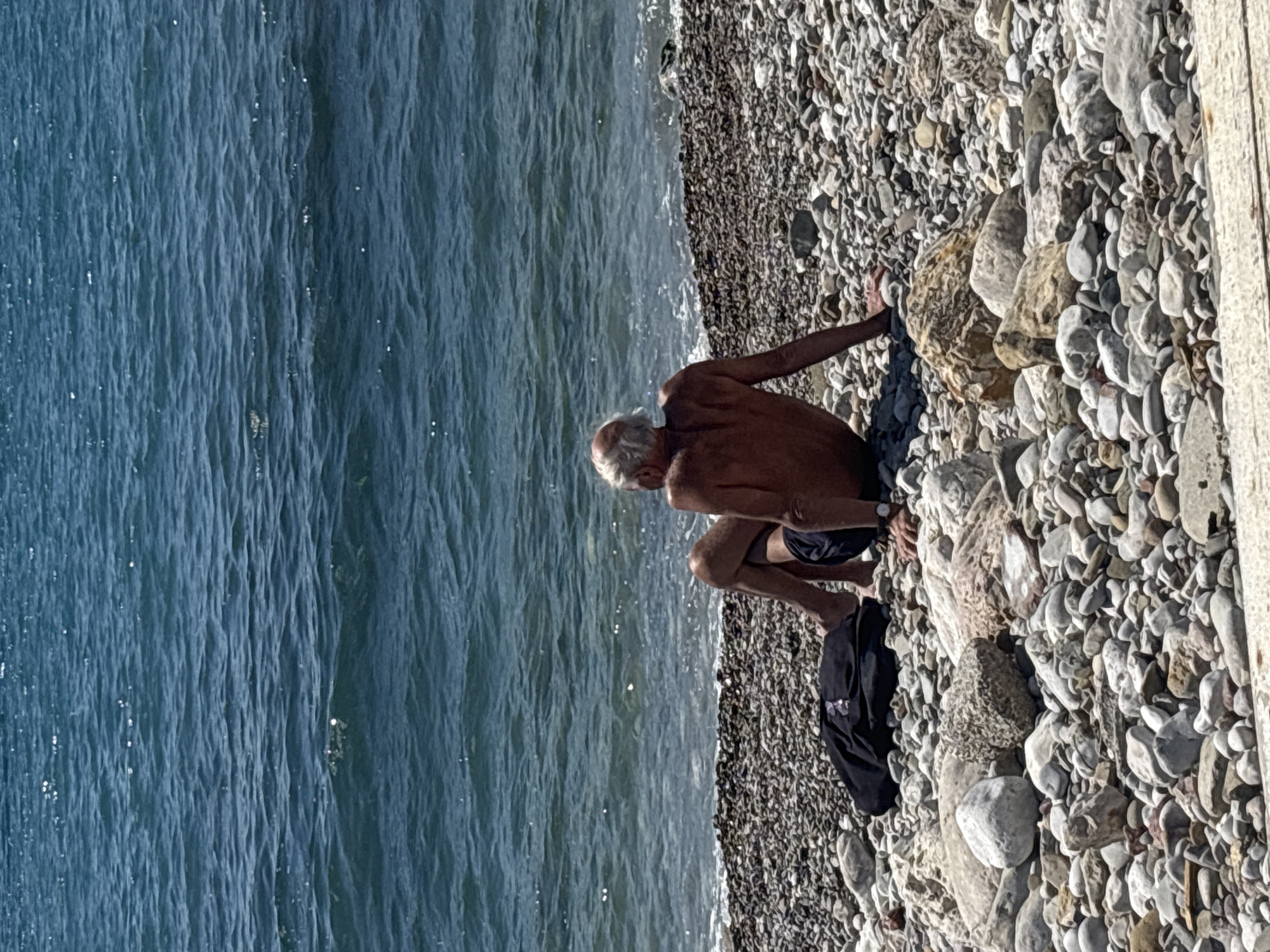 A medium shot captures an elderly, shirtless man crouching on a pebble beach beside the sea. He appears to be interacting with rocks. A dark jacket lies beside him. The location is likely a beach in the city of Bar, Montenegro.