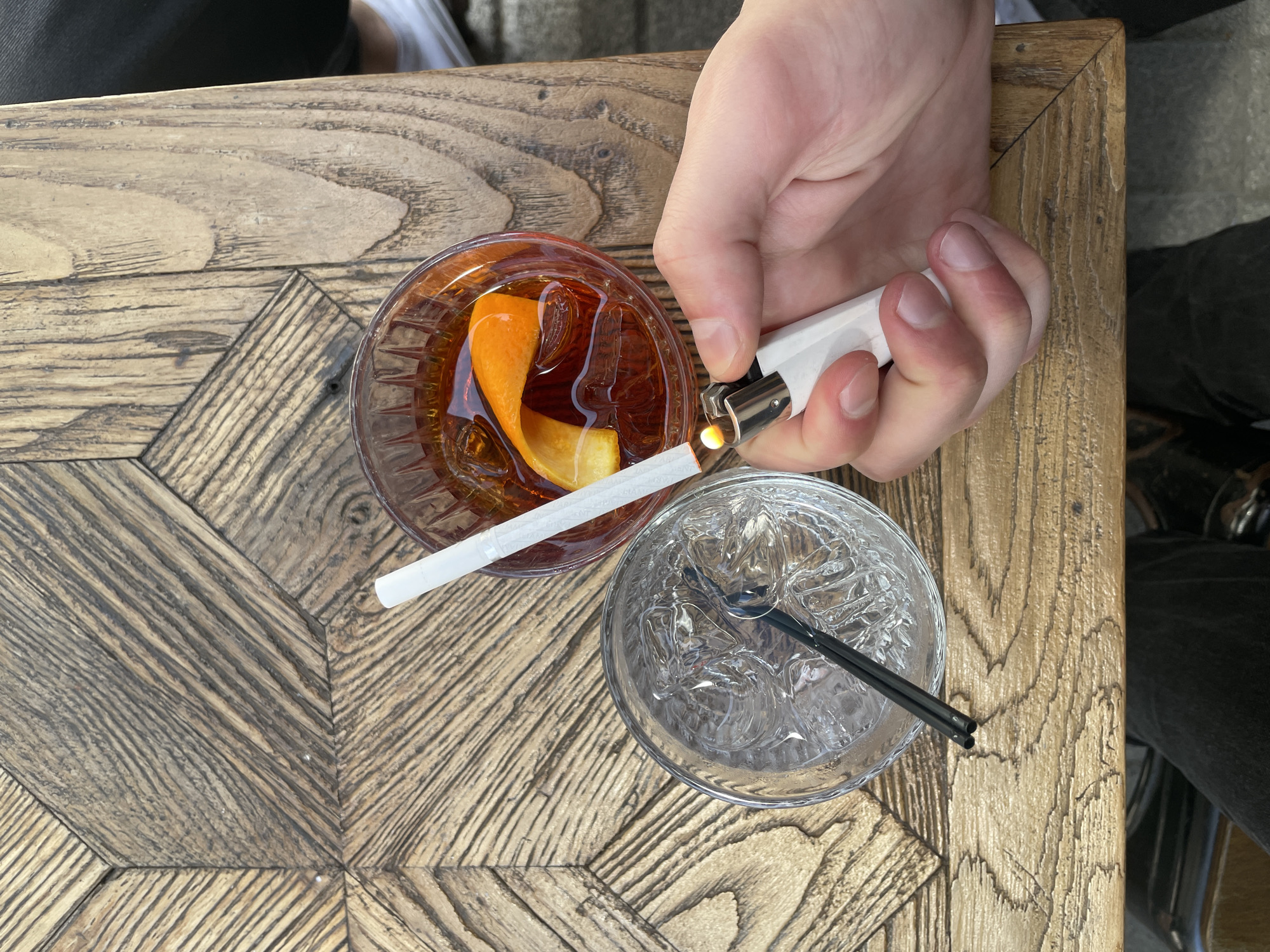 The image shows an overhead view of a wooden table in Kadıköy, Turkey. On the table, there is a glass of amber liquid with an orange peel inside and a glass of water with ice and a black straw. A hand is holding a white lighter and lighting a white cigarette, which rests between the two glasses. The table is made of wood with a patterned design, and part of someone's clothing is visible on either side of the table.