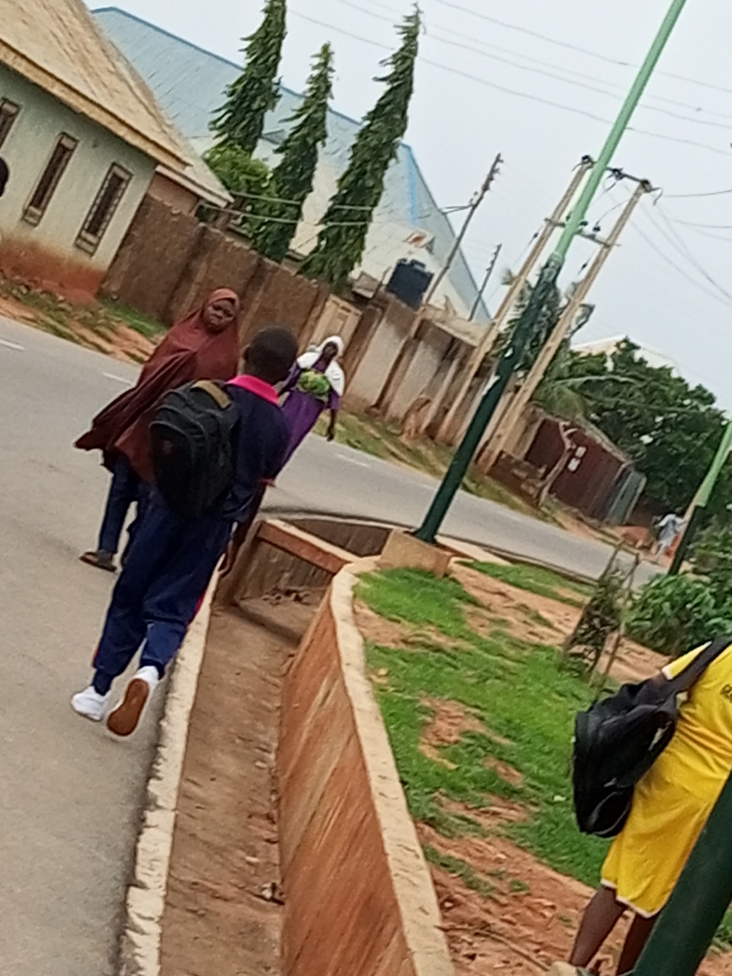 The image shows a street scene in Tempure, Nigeria, with several people and elements of the urban environment. In the foreground, on the right side of the image, a person wearing a bright yellow shirt and dark shorts is seen from behind, carrying a black backpack. Their legs are visible, with one leg bent. To the left and slightly behind this person, a student in a blue uniform with a pink collar and white shoes is walking on a paved walkway next to a drainage ditch. This student is also carrying a black backpack. Further to the left, a person in a dark brown hijab and matching cloak is walking, looking towards the viewer. Another person in a white garment is visible behind the student. The background includes buildings with tiled roofs, fences, trees, and a utility pole with electrical wires. The ground is paved on the walkway and street, with grass and dirt on the sides. The sky is overcast.
