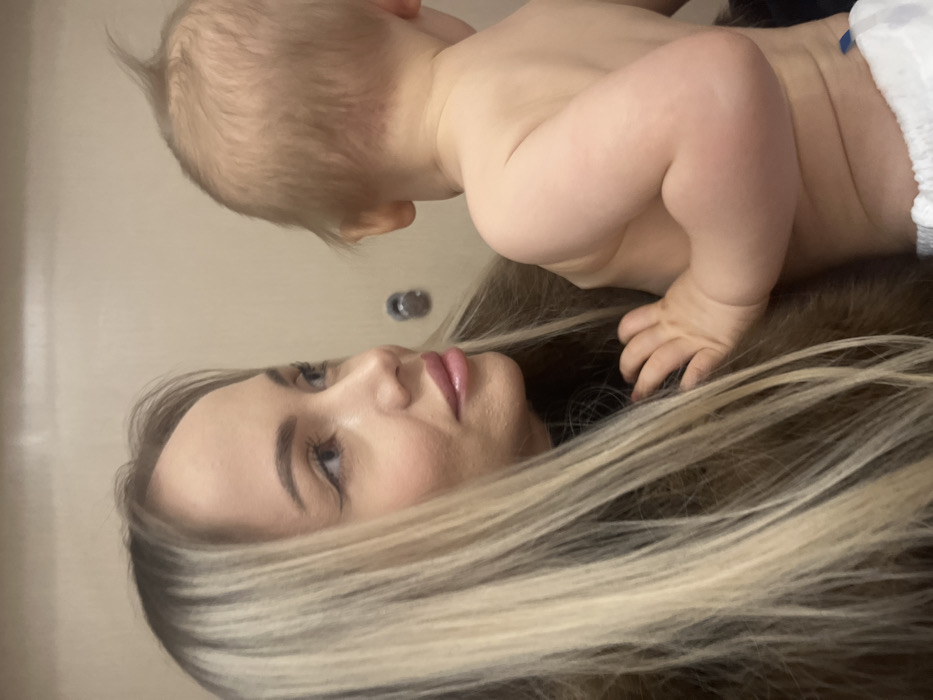 A fair-skinned woman lies on her back with long, blonde-highlighted hair spread beneath her. Her eyes are gazing upwards. A baby in a diaper crawls on top of her hair, with one hand resting on her head. The baby is also fair-skinned and has short blonde hair. A silver knob is visible on the wall behind them. The overall composition is taken from a slightly low angle, emphasizing the faces of both the woman and the child.