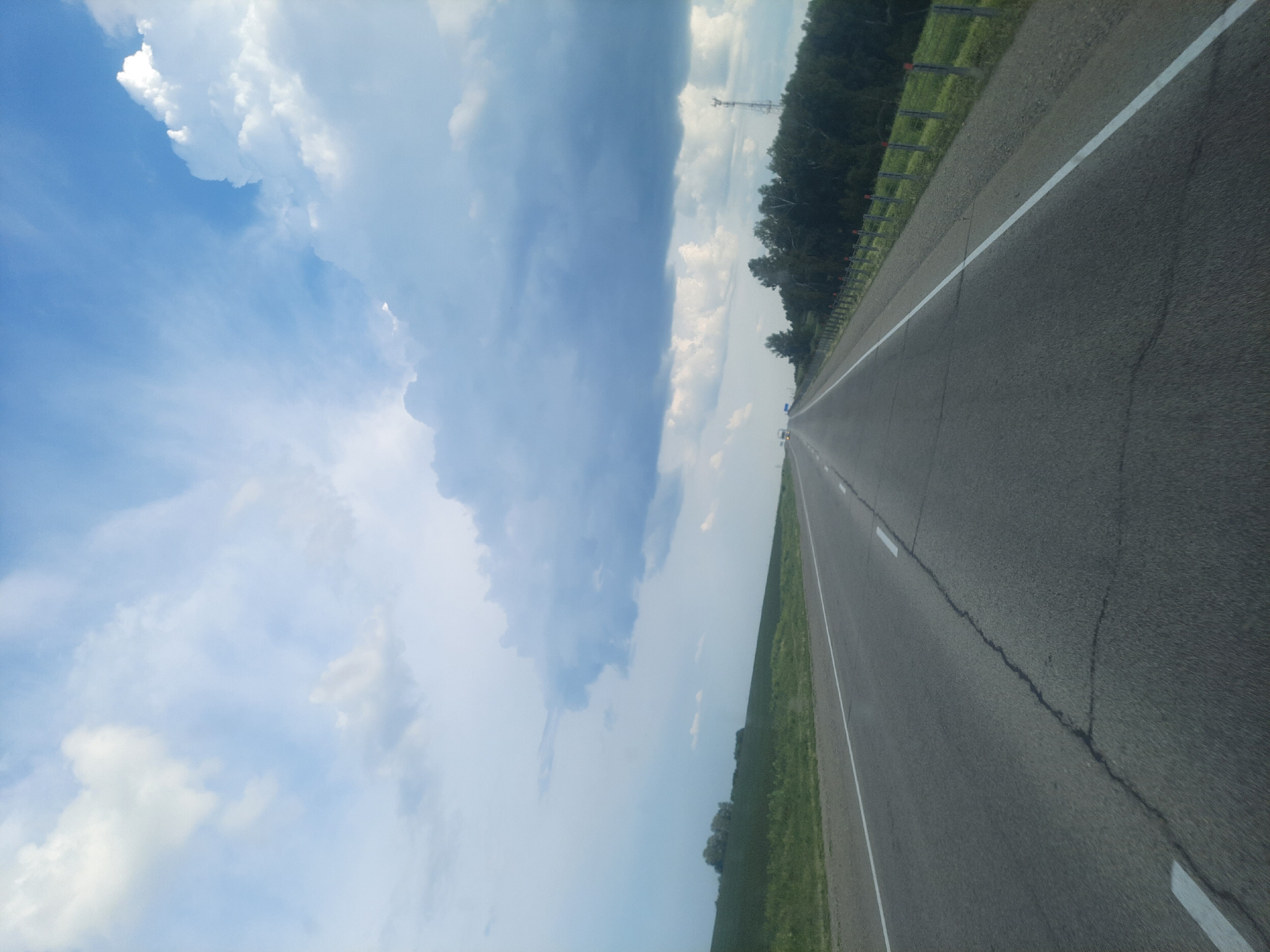 The image shows a view from a moving vehicle on a highway in Oktyabr'skii, Russia. The perspective is from the passenger seat, looking forward and slightly to the right. The road is paved with asphalt and has a white line marking the lanes. The highway is bordered by a grassy shoulder and a fence. To the right of the fence, there is a dense line of trees. The sky is partly cloudy with large, dramatic clouds. The sun is not directly visible, but the sky is bright, suggesting it is daytime, likely mid-afternoon. There is a visible power pylon in the distance on the right side of the frame, behind the trees. The scene conveys a sense of travel and open space. No people are visible, and there is no visible text.