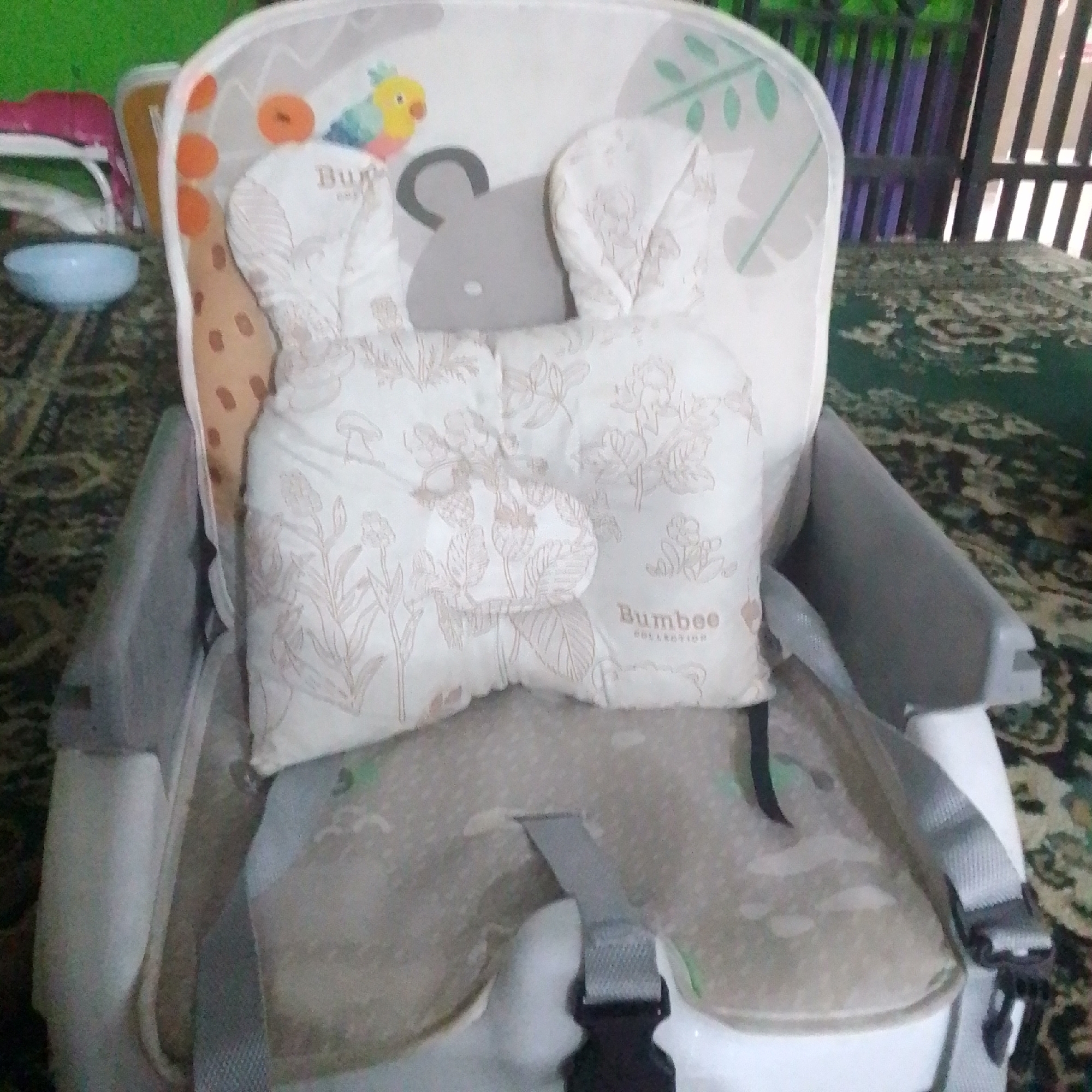In a room in Babakan Jati, Indonesia, a baby high chair is the central focus. The chair is gray and white, featuring a soft white cushion with a subtle floral pattern. A pair of white, plush "bunny ears" are attached to the headrest. The high chair has safety straps, one of which is visible across the seat. The background is blurred, showing hints of green and a patterned rug. A light blue bowl is positioned to the left of the high chair. The overall scene suggests a domestic environment, likely where a baby is fed or plays.