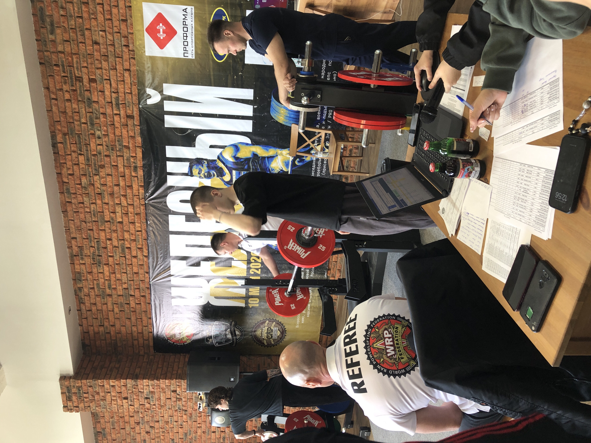 The image shows an indoor powerlifting competition, likely in Chaikovsky, Russia, based on the Cyrillic text and branding. A lifter is preparing for a squat with a barbell loaded with red plates. A "REFEREE" in a white shirt with a WRP (World Raw Powerlifting Federation) logo observes. Behind the lifter is a backdrop featuring advertisements and competition details, including the date "10 MA 202." Another man adjusts the weight on a machine in the background. A table to the side has scoring sheets, a laptop, beverages, a microphone, and personal items, suggesting it's used for recording results. A brick wall is visible in the background. A person at the table is writing on the sheets, apparently documenting the event.