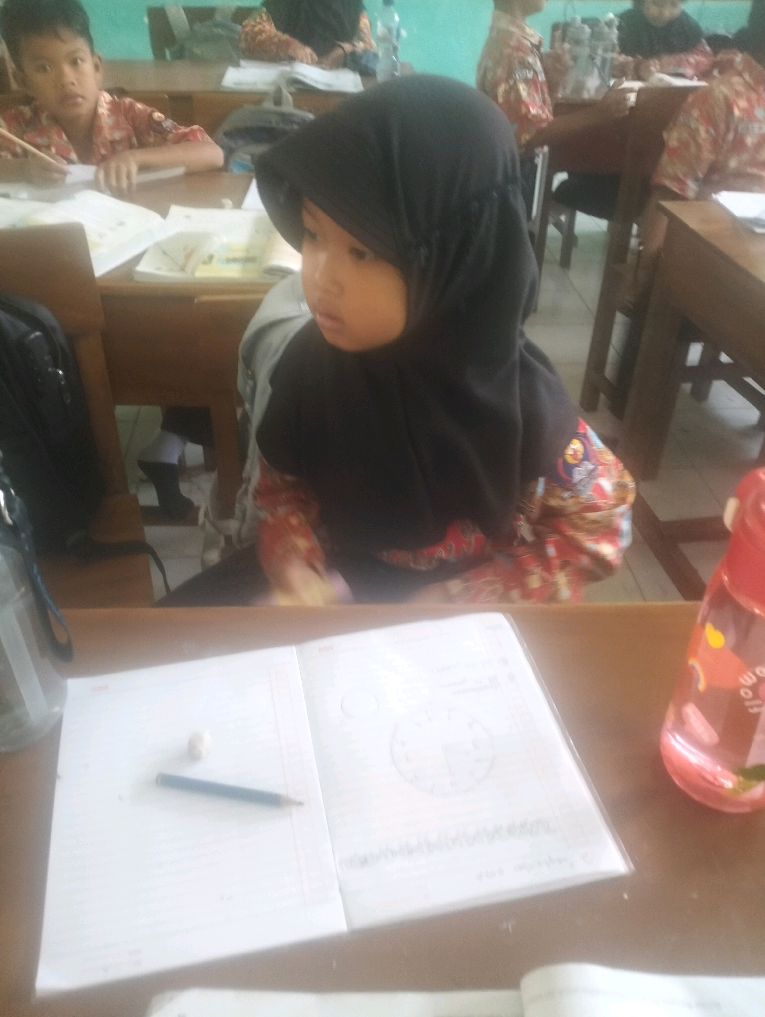 In an Indonesian classroom in Blabakan, a young girl wearing a black hijab and a colorful shirt is seated at a wooden desk. She is looking to her left, and her face is partially obscured by the hijab. In front of her on the desk are an open notebook, a pencil, and an eraser. The notebook appears to be a school assignment, with diagrams and text visible. To the right of the notebook is a pink water bottle with a cartoon character on it. In the background, other students are visible at their desks, as well as classroom furniture and what appears to be a whiteboard. The lighting in the room is natural, coming from windows, and creates a warm, inviting atmosphere. The overall scene depicts a typical day in an Indonesian elementary school.