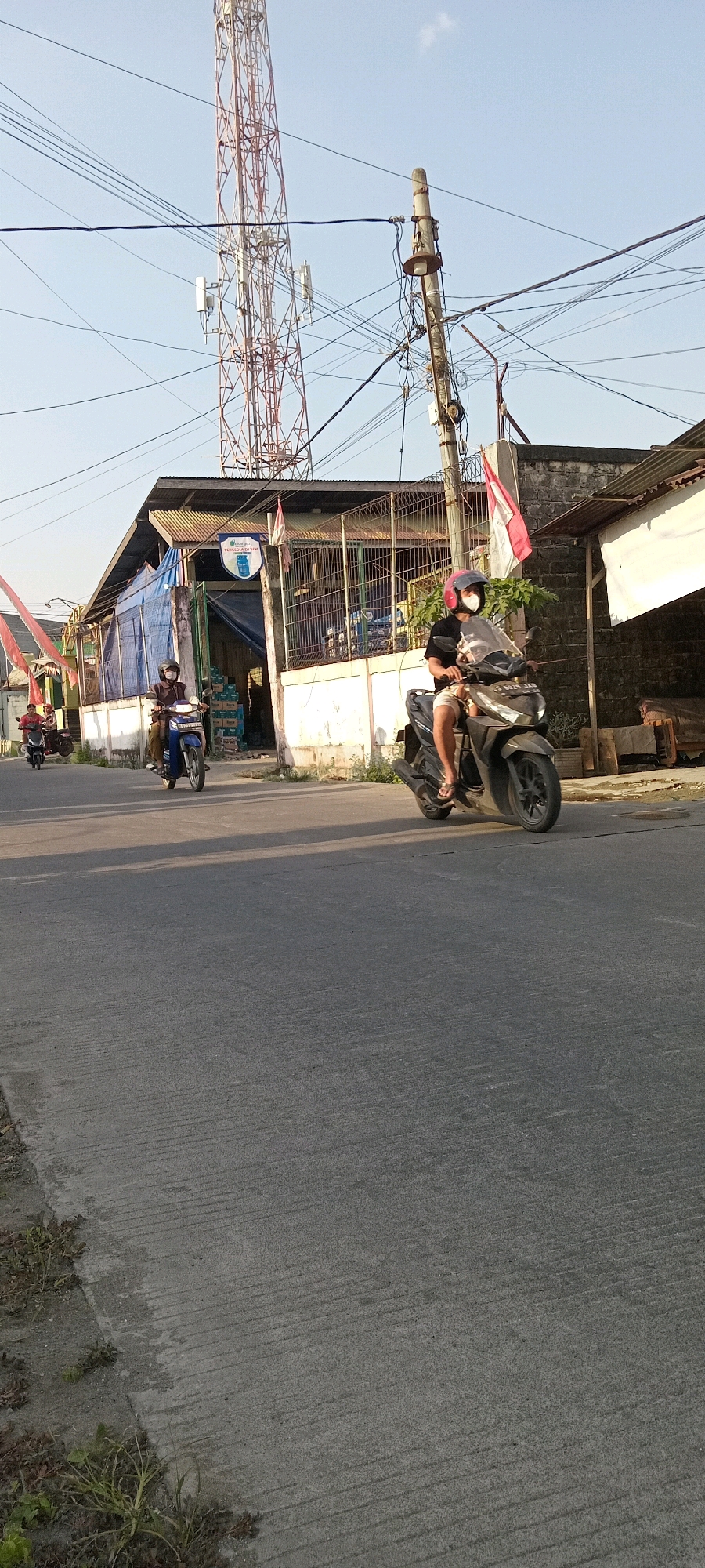 The image shows a street scene in Babat, Indonesia. In the foreground, a gray asphalt road with textured lines runs diagonally across the frame, with sparse vegetation on the left edge. A person on a dark-colored scooter is riding towards the right side of the image, wearing a helmet and shorts. Behind this scooter, a person on a blue scooter is also moving towards the right, wearing a helmet and visible through the scooter's windshield. To the left of the road, a building with a corrugated metal roof and white walls is visible. A tall, lattice cell tower stands behind the building, accompanied by numerous power lines and a concrete utility pole with a light fixture. A red and white Indonesian flag is attached to the building. In the distance, a smaller cell tower is also visible. The sky is clear and blue.