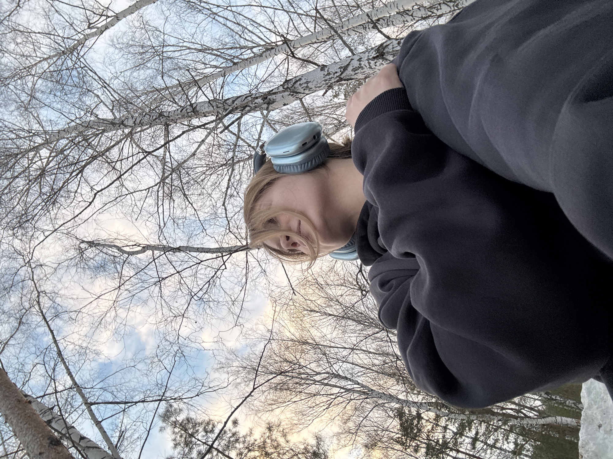A low-angle outdoor image depicts an individual in a wooded area in the city of Noviy, Russia. The subject, a person with light-colored hair, is visible from the upper torso, wearing light blue over-ear headphones and a dark grey or black oversized hoodie. Their head is tilted upwards and slightly to the right. The person's right arm is bent, with the hand resting on their left arm. The background consists primarily of numerous leafless birch trees with white bark, extending upwards to a partially cloudy sky displaying both blue and faint yellowish hues. Patches of snow are visible on the ground in the lower-left quadrant. Some evergreen trees are also discernible on the far left of the frame.