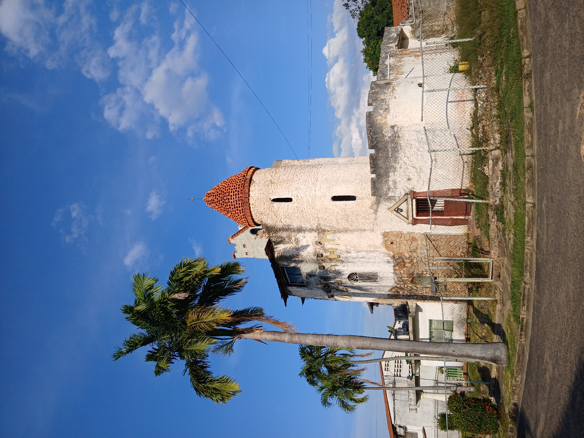 The image presents a partially obscured view of the Old Customs building located in Guayana City, Venezuela. The old customs building has a white exterior, cylindrical tower with a reddish-orange tiled roof. The tower has a few small, rectangular window openings. The building is aging, showing signs of decay and weathering.
A palm tree occupies the foreground, partially obscuring the lower portion of the building and partially casting a shadow on it. Other modern buildings are visible behind the palm trees.
The surroundings consist of a street with an adjoining sidewalk. There is a chain-link fence along the street separating the building from the road. The sky above is a clear blue, scattered with fluffy white clouds. A power line stretches across the sky, intersecting the top portion of the building.
