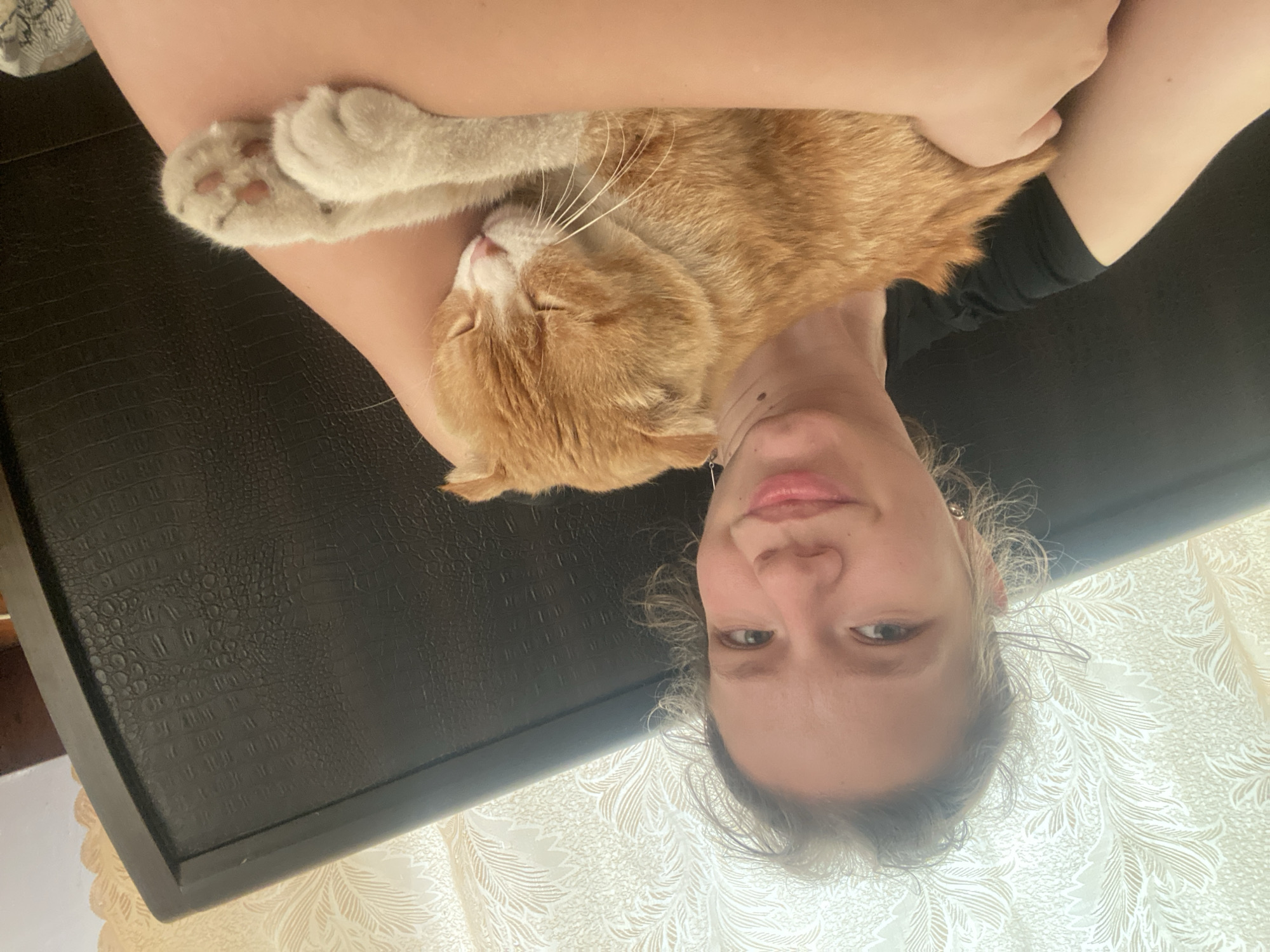 An upside-down close-up shot depicts a woman holding an orange tabby cat, apparently indoors. The woman's face is visible, with light skin and light-colored hair. The cat is nestled in her arms and appears to be sleeping. The surface beneath them is a dark, patterned material. The location of Pakhynia, Ukraine is not ascertainable.