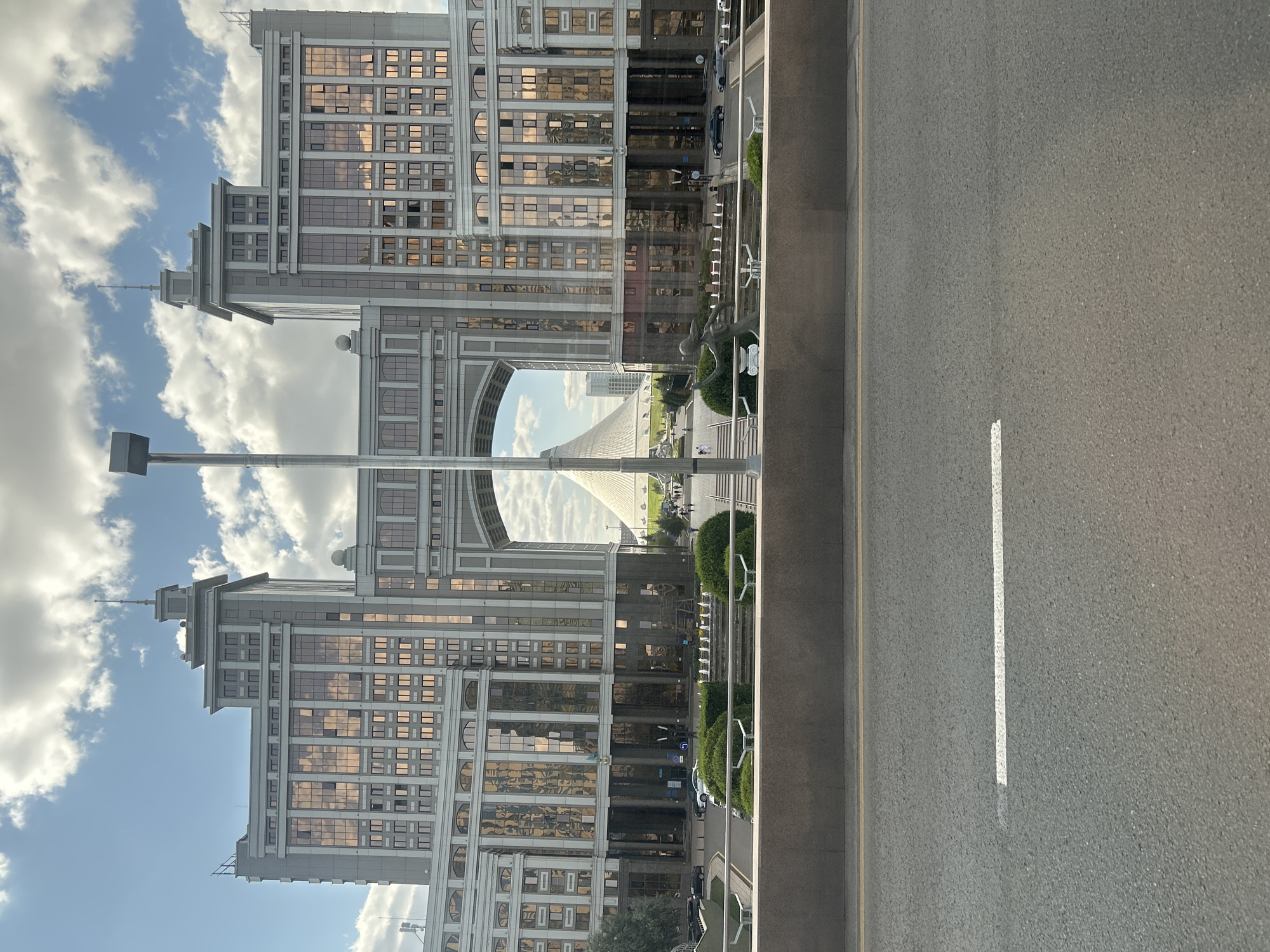 The image shows a wide shot of a grand archway connecting two tall, modern buildings with reflective glass facades in Astana, Kazakhstan. The archway frames a view of a unique, conical-shaped building in the distance, possibly a cultural landmark. The buildings flanking the archway are symmetrical and feature a mix of grey stone and gold-tinted windows, reflecting the cloudy sky. In the foreground, a concrete barrier separates the viewpoint from a road with a white lane marking. Between the barrier and the buildings, a low wall with decorative elements and a few people walking can be seen, as well as some sculpted bushes and a lamp post. The scene appears to be an urban setting on a bright, partly cloudy day.