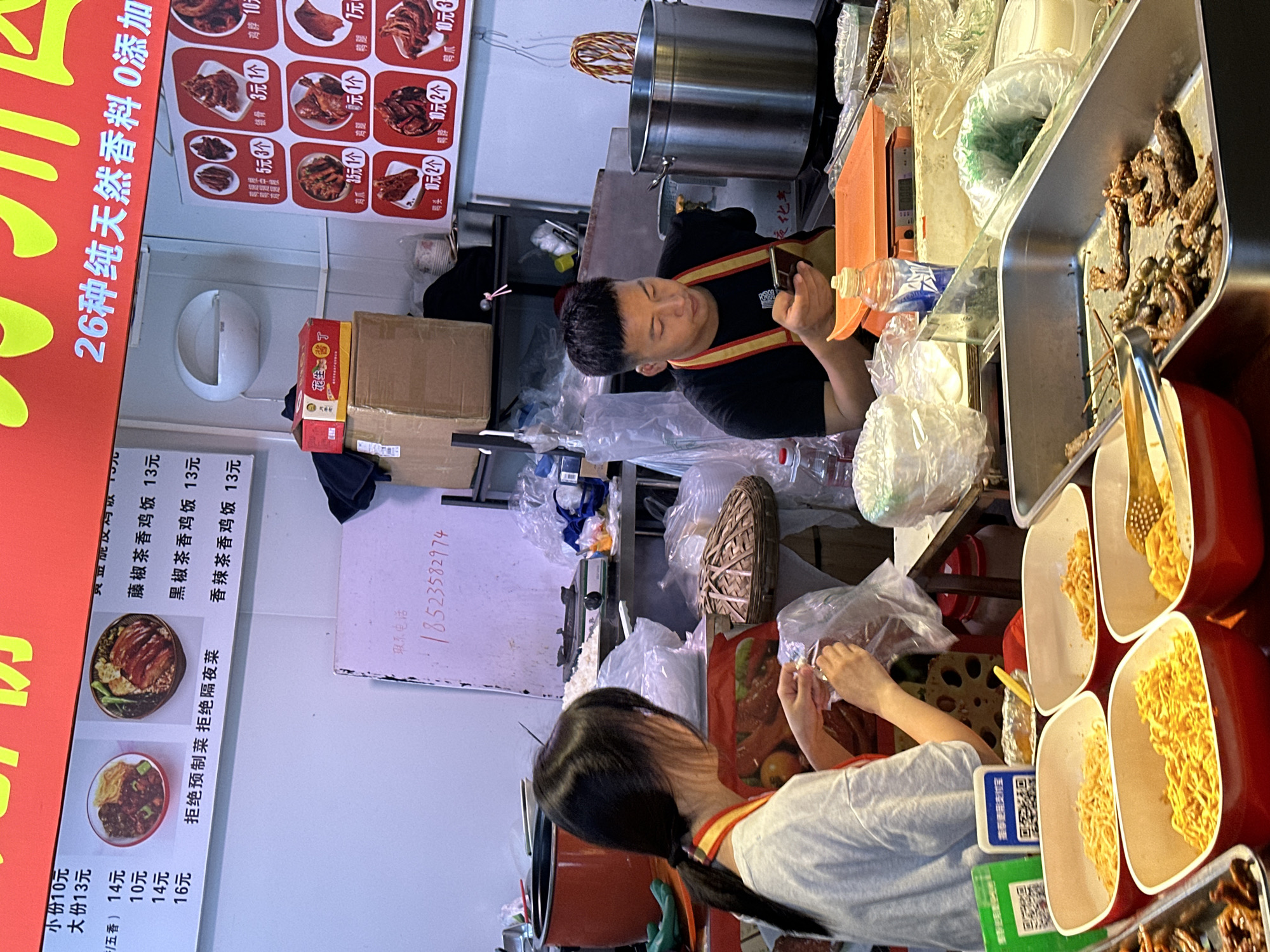 The image depicts a food stall in Jiangjin, China. A man is looking at his phone behind the food counter while wearing a black shirt with orange and yellow straps. A woman in a grey shirt with orange and yellow straps is in the front of the counter making food. Food items like noodles, meat, and peanuts are on display. Signage indicates the stall offers dishes like "pepper tea chicken" and "spicy tea chicken." A telephone number is visible on a whiteboard. QR codes suggest mobile payments are accepted.