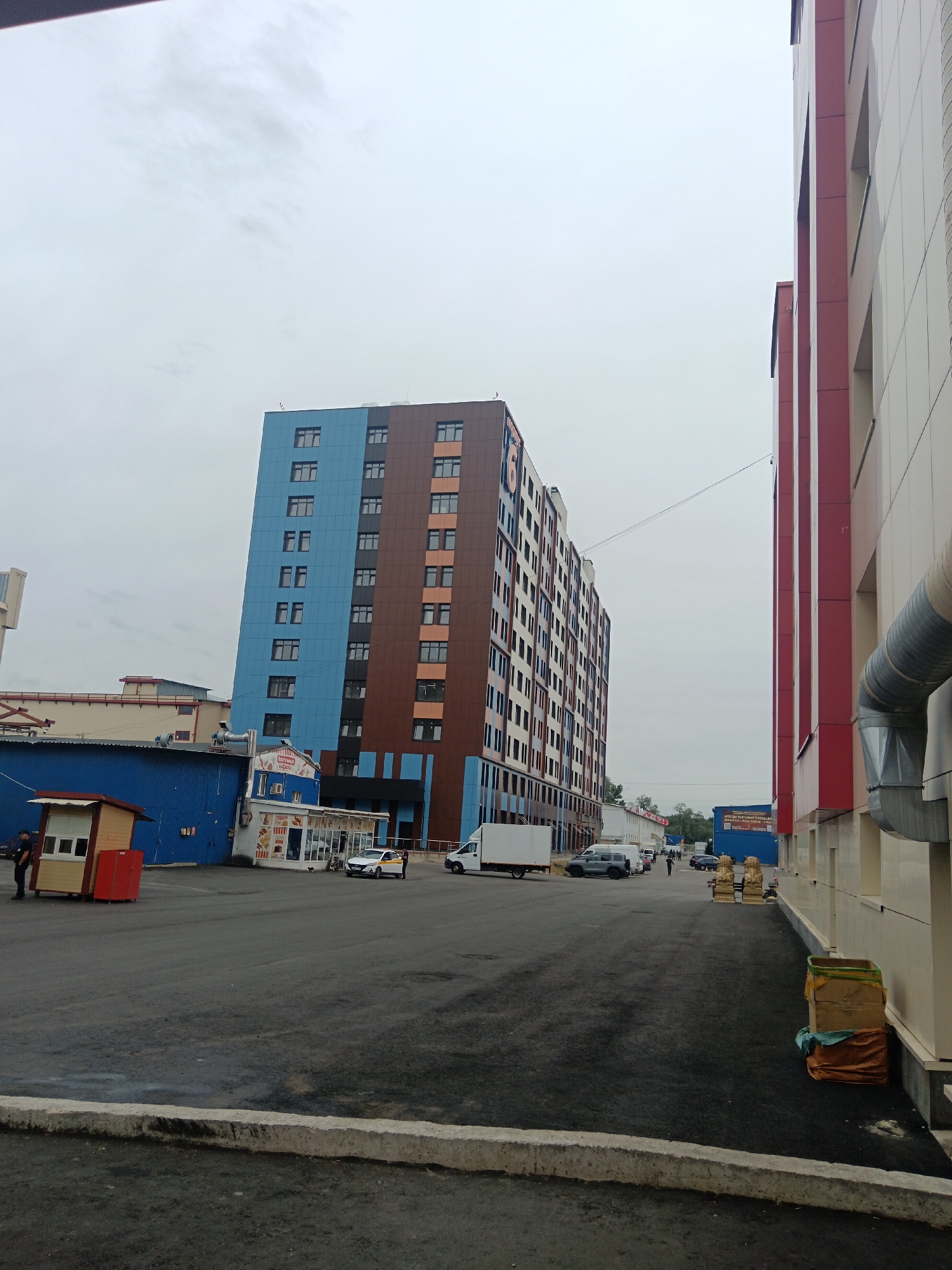 An outdoor medium shot captures a street scene with a tall, multi-story building dominating the left side of the frame. The building is clad in alternating sections of blue and brown panels and has numerous windows. To the right of this building is a smaller, blue, single-story structure with a white facade and signage. Adjacent to the blue structure is a small, wooden kiosk with a red base. Several vehicles are present: a white van is parked near the blue building, and a white car is driving away. A dark-colored SUV is parked in the middle distance. Further down the street, other buildings and vehicles are visible. The ground is paved with asphalt. The sky is overcast, and the overall lighting suggests a cloudy day.