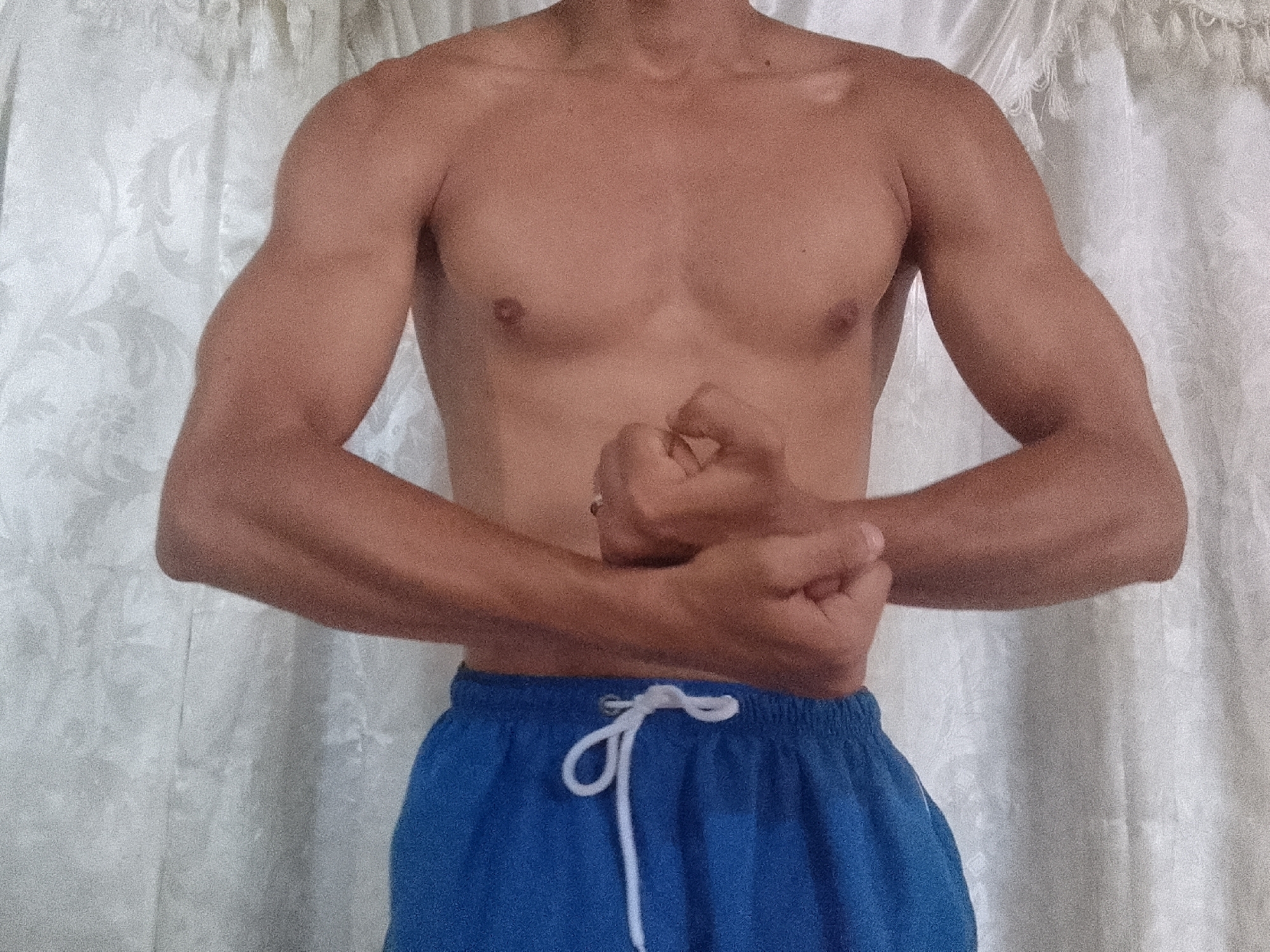 A shirtless man with a light tan is posing indoors, flexing his arms and chest. He is wearing blue shorts with a white drawstring. The background is a light-colored, textured curtain or sheet. The location is not discernible in the image; there is no information to indicate that this photo was taken in Juan Griego, Venezuela.