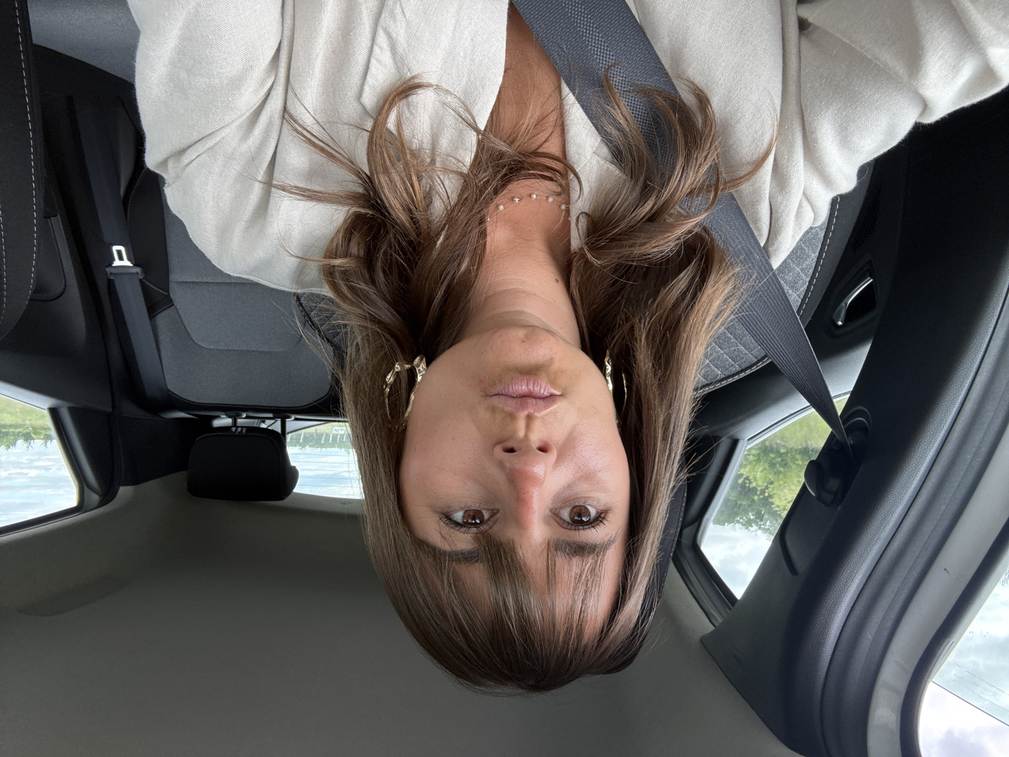A person is seated inside a car, wearing a seatbelt. The person has brown hair with bangs, and is wearing gold hoop earrings and a white pearl necklace. The seat's upholstery is grey. A body of water and green vegetation can be seen through the car window. The photograph was taken at an upside-down angle. The location is Sury-le-Comtal, France.