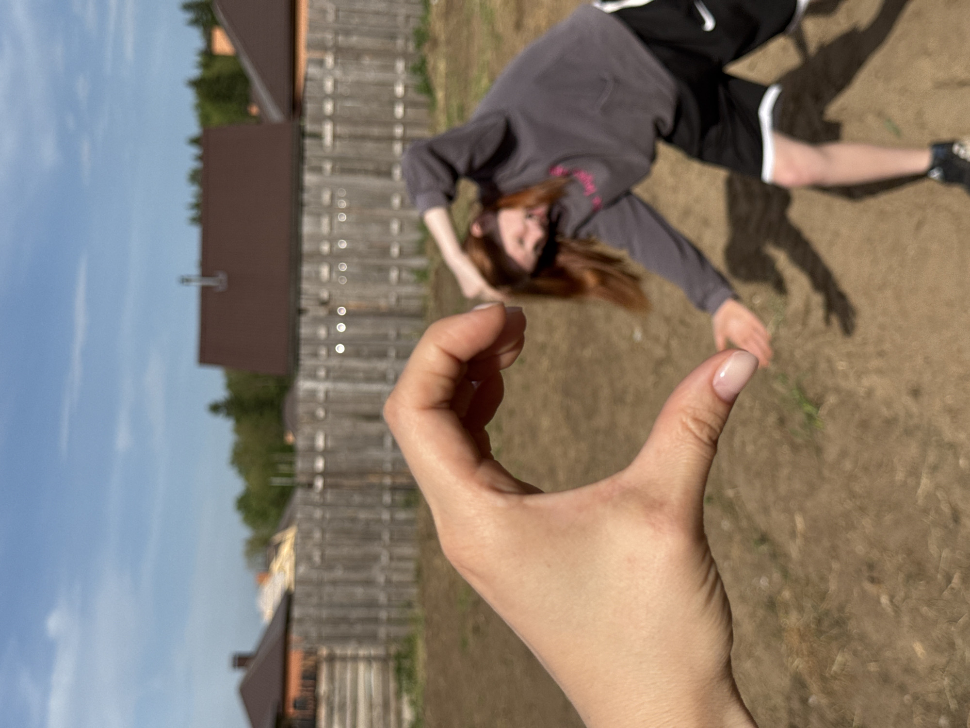 A person is lying on the ground outside in Chaikovsky, Russia. A hand is raised in the foreground. There is a wooden fence and several houses in the background. The sky is blue.