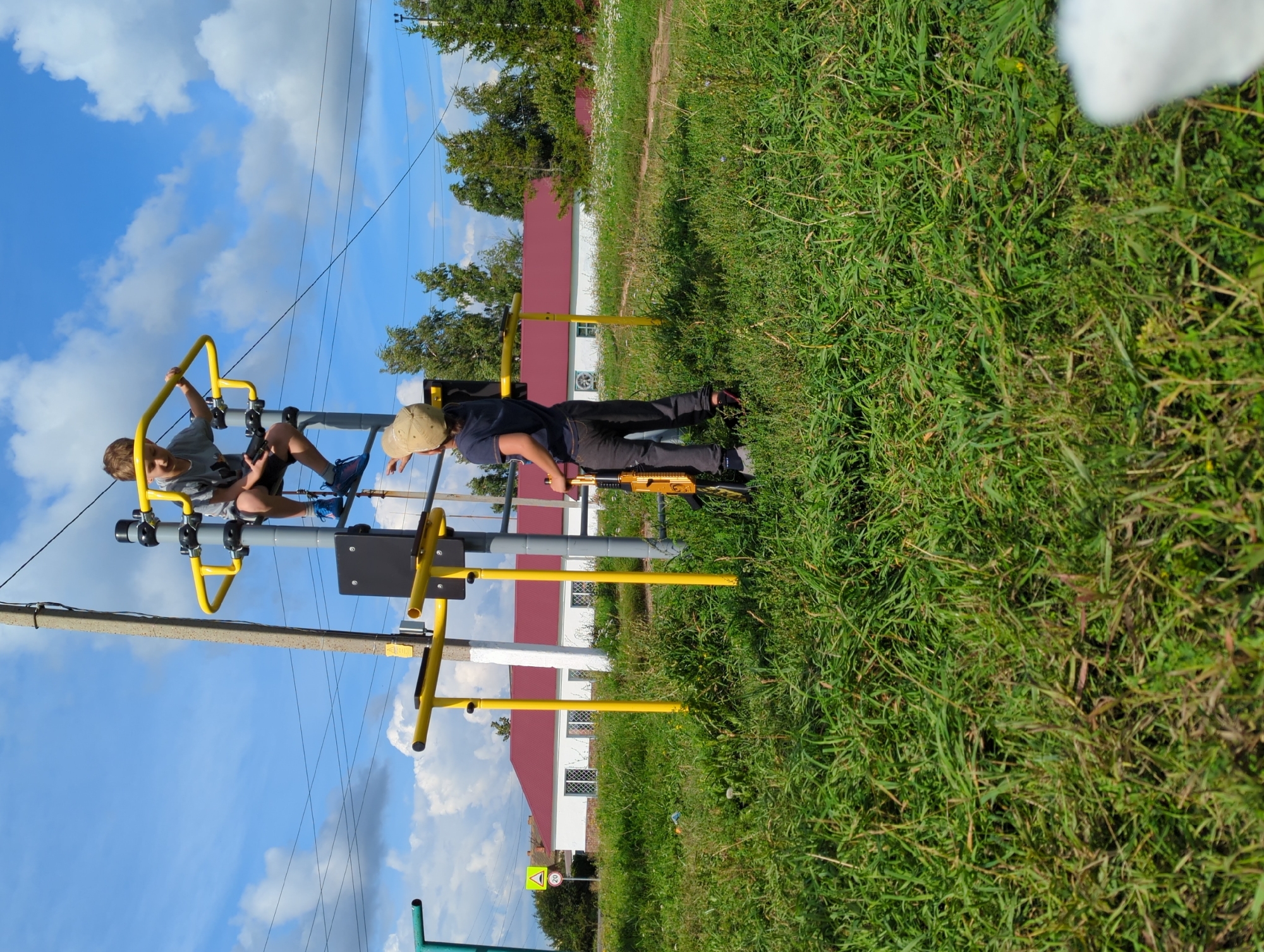 The image depicts an outdoor scene in Tenishevo, Russia, featuring two young boys engaged with a yellow outdoor exercise apparatus. The boy on top of the apparatus, wearing a grey t-shirt and blue sneakers, is looking downwards. The second boy, wearing a beige cap and a dark blue t-shirt, stands below, holding a golden toy gun and looking up at the first boy. The exercise equipment is positioned on a grassy area with a white building with a red roof in the background. A road sign indicating a speed limit of 20 is visible to the left. The sky is partly cloudy.
