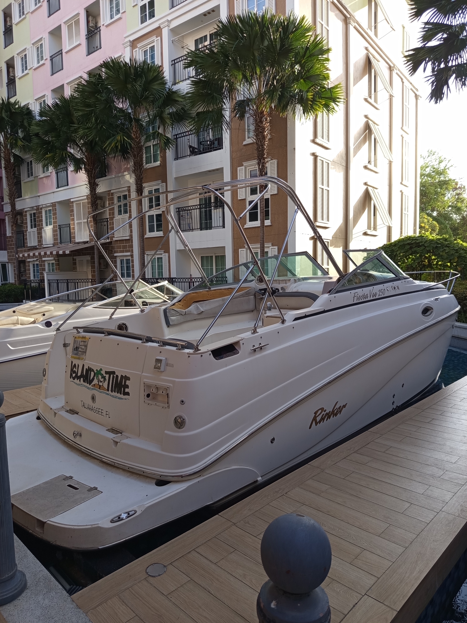 The image shows two white boats docked at a wooden pier. The boat in the foreground has "ISLAND TIME" written on its stern, along with "TALLAHASSEE, FL". The boat also has "Fiesta Vee 250" and "Rinker" written on it. Behind the boats is a multi-story building with pastel-colored facades and numerous windows with white shutters. Palm trees are visible in front of the building and between the boats. The scene appears to be a marina or waterfront area within Pattaya City, Thailand, given the presence of tropical vegetation and architecture commonly found in such locations. The lighting suggests it is daytime, with sunlight casting shadows. There are no people visible in the image.