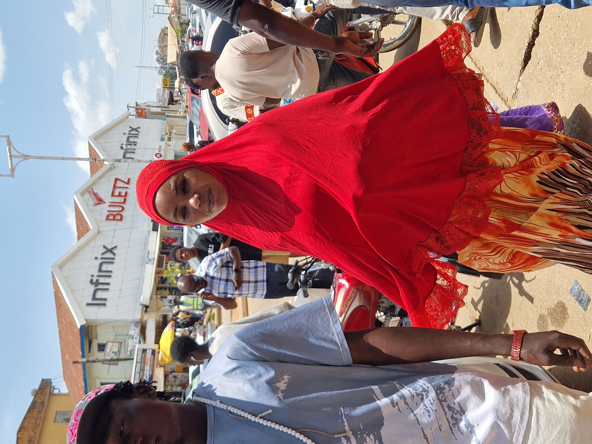 In a street scene in Unknown, Unknown, a group of people are seen outdoors under a bright, cloudy sky. In the foreground, a woman wearing a vibrant red hijab and a patterned dress stands prominently, facing forward with a neutral expression. To her left, a man in a light blue t-shirt and a baseball cap is partially visible, wearing a long beaded necklace. Behind them, other individuals are scattered, including a man in a plaid shirt talking on a phone and others walking or standing near parked vehicles. Several motorcycles are also present, one with a red frame visible in the mid-ground. The background features buildings with signage, including "Infinix" and "BULETZ," suggesting a commercial area. The ground is paved with asphalt, showing some cracks and debris. The overall atmosphere appears to be that of a bustling urban environment.