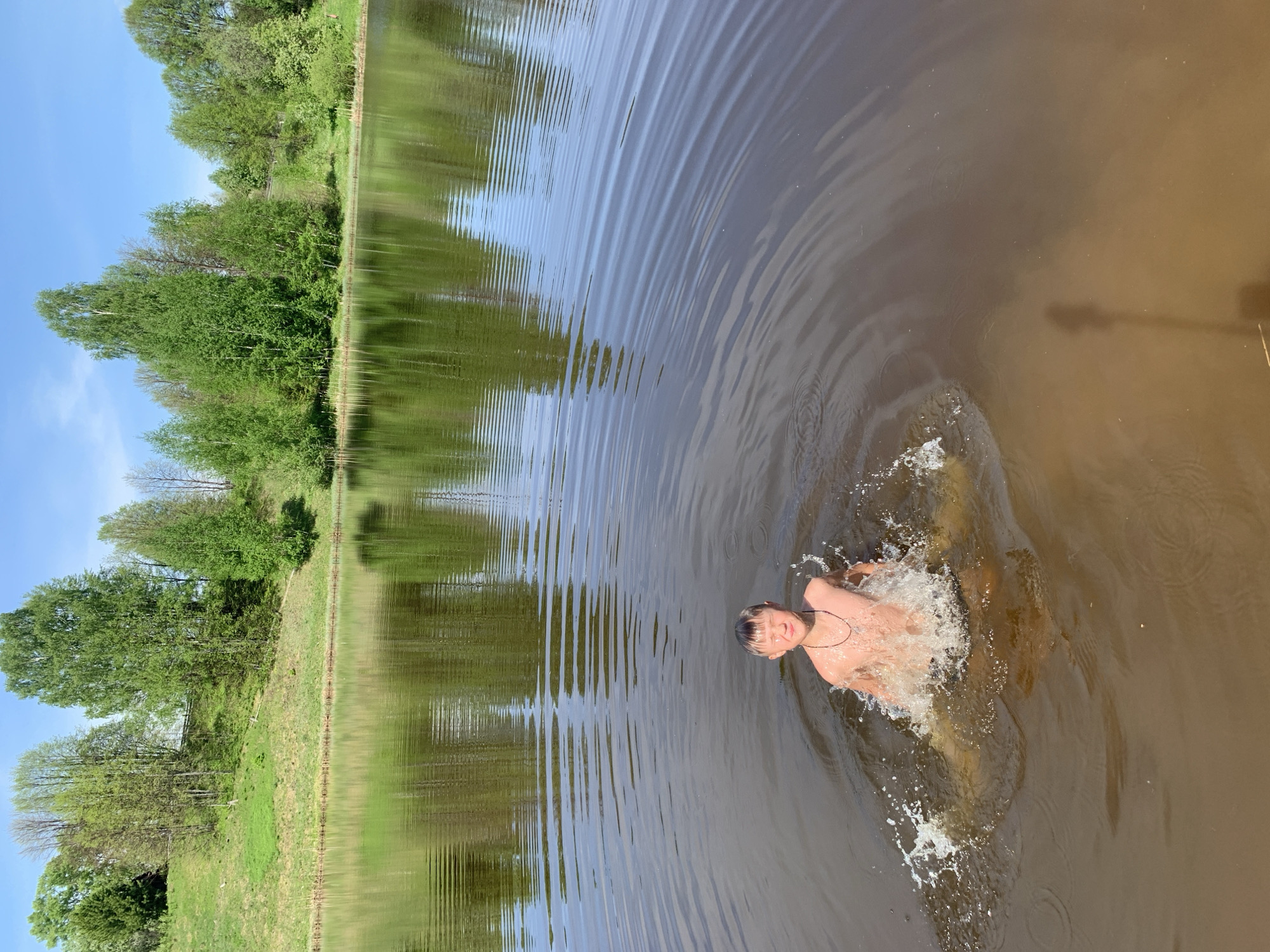 A fair-skinned male, shirtless, is swimming in a body of water. The water is murky and dark, with noticeable ripples and splashes around the individual. The surrounding environment includes green vegetation along the shore and a bright sky visible above the trees. The location is in Zasechnyi, Russia.