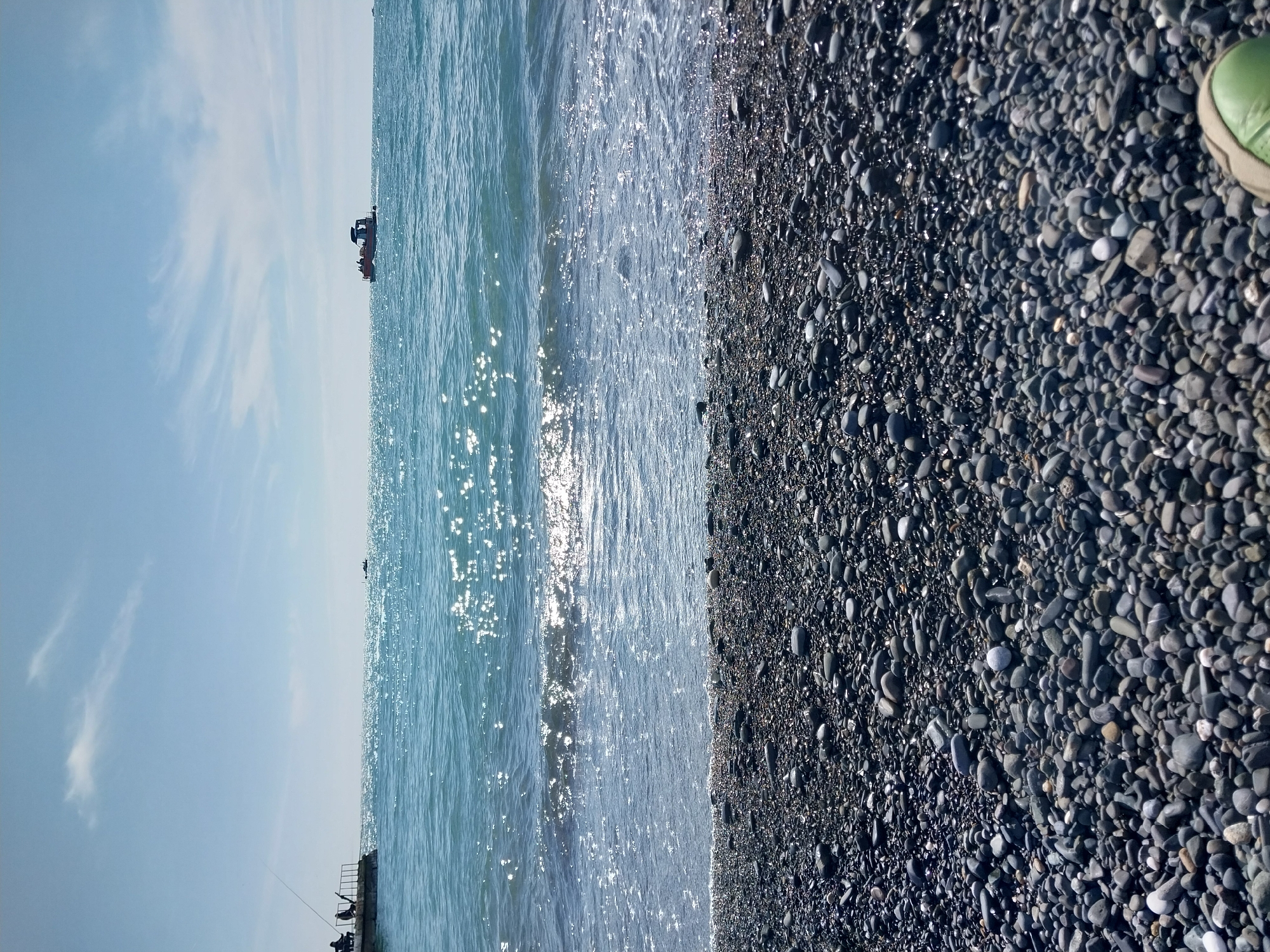 This image captures a serene coastal scene at a pebble beach, likely in Sochi, Russia, given the provided context. The picture is oriented vertically but depicts a horizontal landscape.
The upper portion of the frame features a clear, light blue sky with sparse, wispy white clouds, suggesting fair weather. Below the sky, the vast expanse of the sea stretches out, characterized by calm, blue-green water. Numerous sparkling reflections from the sun create bright, shimmering patches across the water's surface, particularly in the middle distance. Two small boats are visible on the water: one closer to the left, appearing to be a small recreational or fishing vessel, and another smaller boat further in the distance. On the far left, a dark pier or structure extends into the water, with two figures silhouetted against the bright sea, actively engaged in fishing with rods cast into the water.
The right side and foreground of the image are dominated by a dark pebble beach, composed of countless grey, black, and some lighter-colored rounded stones of varying sizes. The texture of the pebbles is clearly visible, some appearing wet from the receding tide. In the bottom right corner, a portion of a bright green shoe or sandal is visible on the pebbles, indicating the perspective of someone standing on the beach.
The scene conveys a peaceful atmosphere, typical of a sunny day by the sea. The time of day appears to be daytime, possibly late morning or early afternoon, due to the bright sun. No visible text is present in the image.