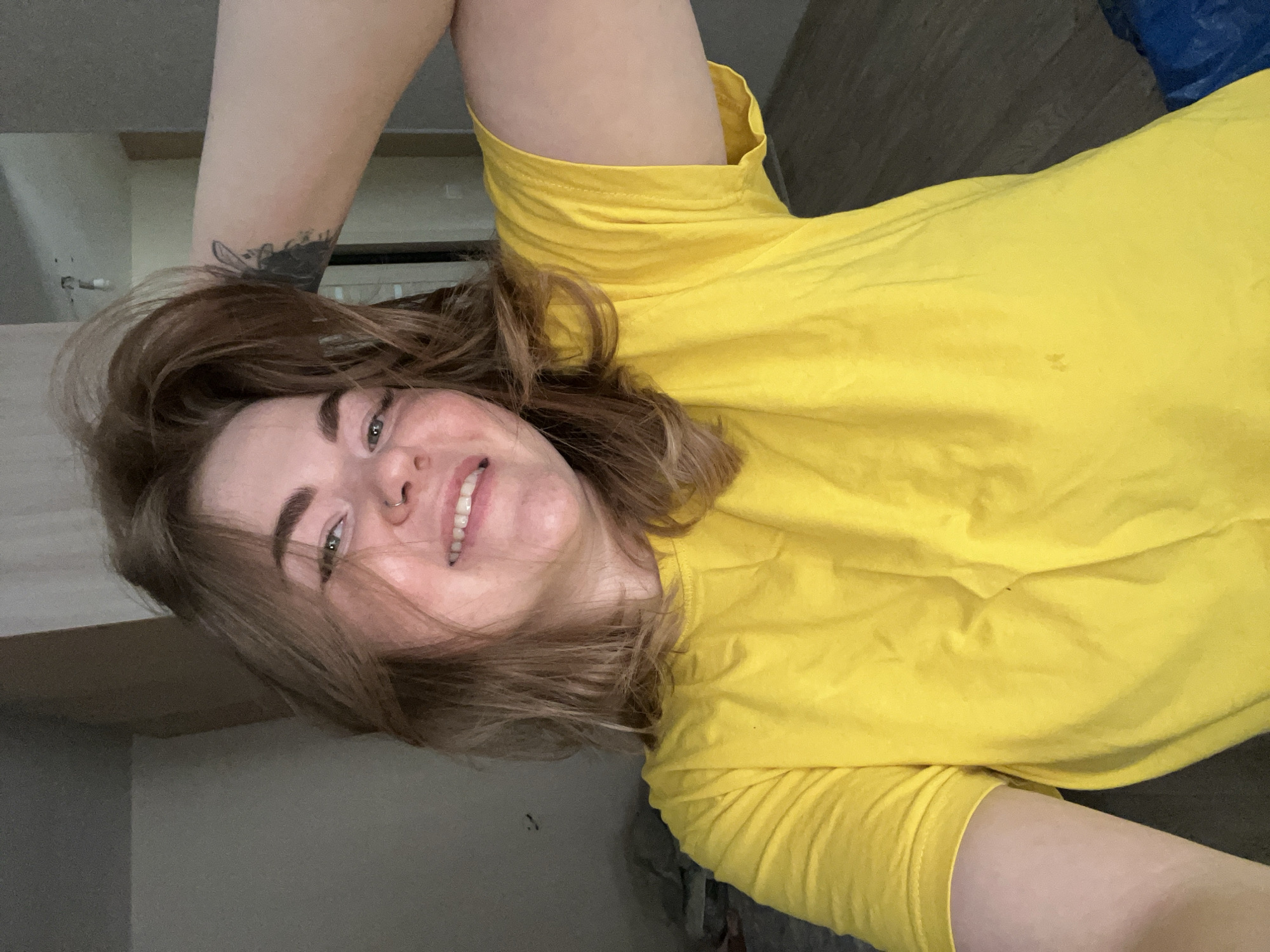 A young woman with fair skin, brown hair, and a nose ring is smiling at the camera. She is wearing a yellow t-shirt and has a tattoo on her arm. The background is blurry but appears to be a room with a cabinet and a wooden floor. The location of Chaikovsky, Russia, is not discernible from the image.