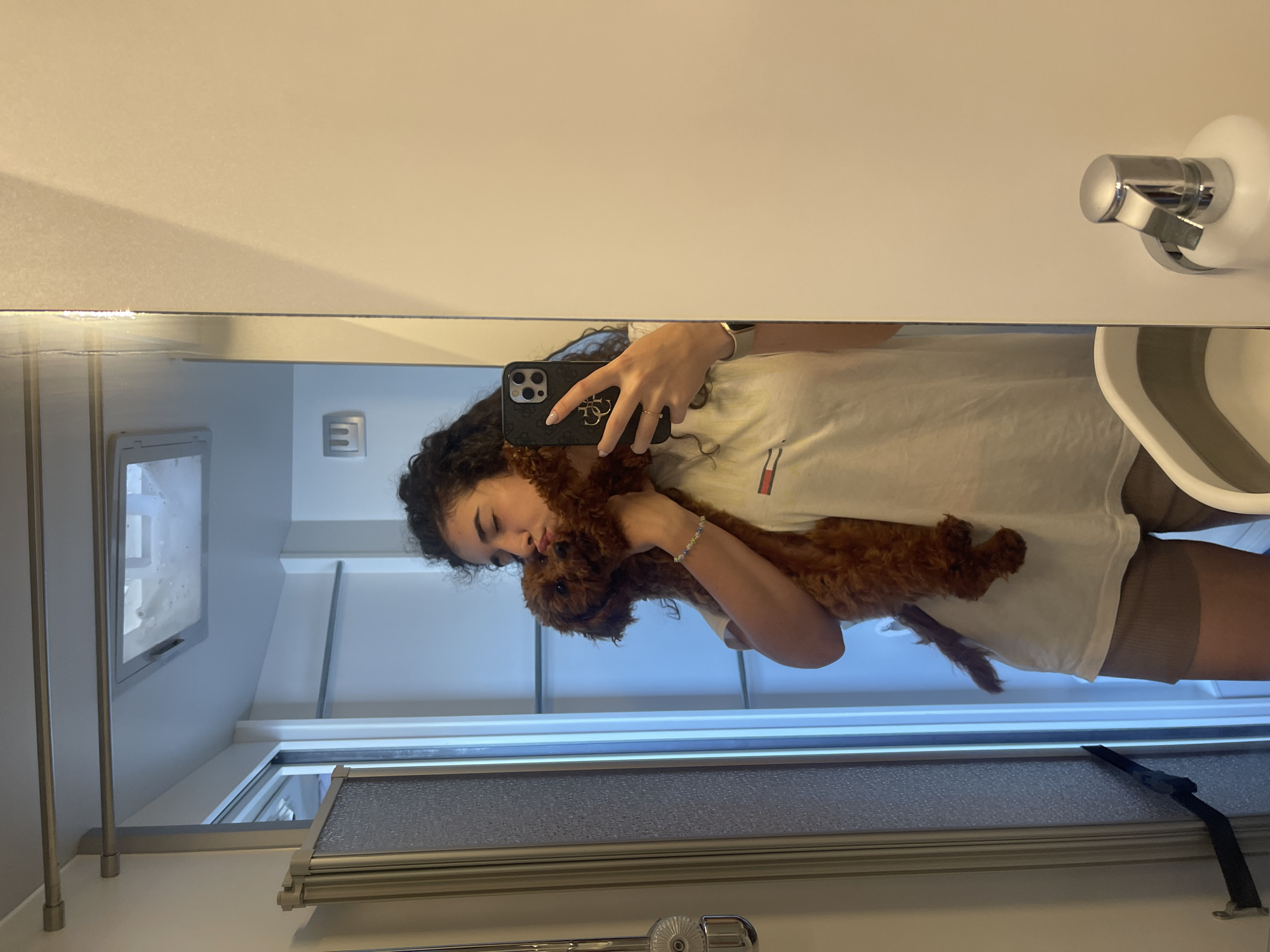 The image depicts a young woman in a selfie reflected in a large mirror, located in Koprinka, Bulgaria. She is holding a small, reddish-brown, fluffy dog against her face, with her eyes closed, appearing to kiss or nuzzle it.
The woman has dark, curly hair and is wearing a light-colored short-sleeved t-shirt with a small rectangular logo on the left chest (featuring red, white, and blue stripes). She is also wearing light brown ribbed shorts. On her right hand, she holds a black smartphone (an iPhone with a multi-lens camera array) in a case with a prominent gold-colored 'Guess' logo, positioned to take the photograph. Her fingernails are painted a light color, and a thin ring is visible on her left ring finger. On her left wrist, she wears a light-colored smartwatch and a beaded bracelet with green, blue, and white beads.
The reflected environment appears to be a compact, modern interior space, possibly a bathroom or a compartment within an RV or camper van, characterized by white or light-colored walls. To the left of the reflection, a metallic-framed shower enclosure with textured translucent panels is visible, along with a chrome shower head and control mechanism. A black strap or restraint is affixed to the wall adjacent to the shower door.
Above the woman's head in the reflection, two horizontal metallic bars are mounted near the ceiling. Above these bars, a rectangular ceiling vent or small skylight is visible, showing some condensation or texture. The wall behind the woman and dog in the reflection is white, featuring a small square vent or fixture, and another horizontal metallic bar further back. In the lower right of the reflection, a wall-mounted chrome soap dispenser is visible, alongside the partial view of a white collapsible basin or bucket with a gray rim. The entire scene is brightly illuminated.