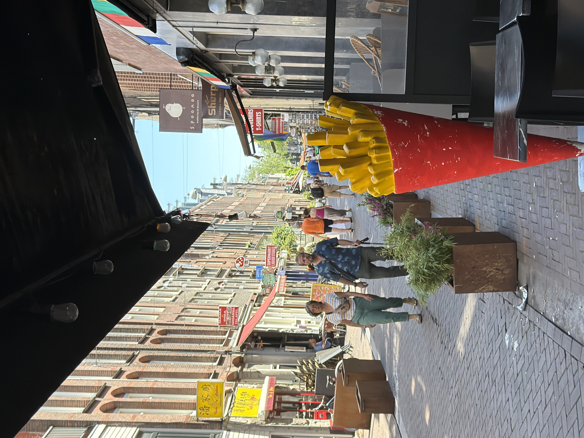 This is a street-level, eye-level shot of a commercial street in Amsterdam, Netherlands, on a sunny day. The perspective is looking down a street lined with multi-story brick buildings, typical of Dutch architecture, with arched windows on the upper floors. The street is paved with cobblestones and has planters with greenery interspersed along the sides.
In the foreground, a large, red cone with yellow french fries is prominently displayed, likely a decorative item for a food establishment. To the left of the fries sculpture, two people, a man and a woman, are walking towards the camera. The woman is wearing a striped top and green pants, and the man is in a patterned blue shirt and dark pants. They are walking on the cobblestone pavement.
Further down the street, several people are walking, and some are seated at tables outside a cafe. Various signs are visible on the buildings, including those for "SPOONOO" (bubble tea & dessert), "SHERPA", "SOUVENIRS T-SHIRTS", and "SUPERMARKT LIQUOR STORE", indicating a mix of retail and food businesses. An awning covers part of the sidewalk in the foreground. The sky is clear and blue. A trash can is visible on the left side of the street.