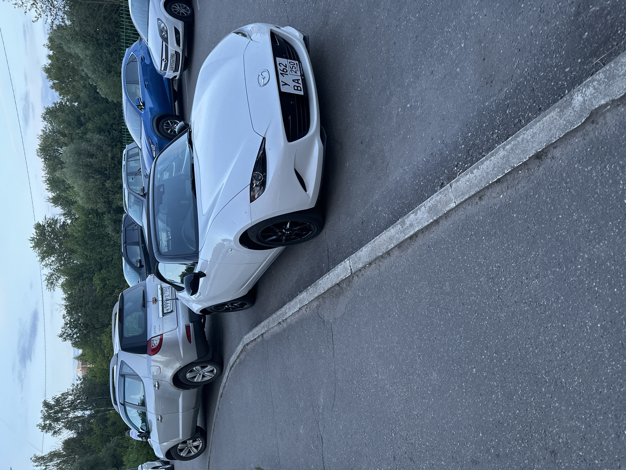 The image captures a row of parked cars on an asphalt surface next to a concrete curb, with a line of trees bordering the area. The scene appears to be an outdoor parking lot or street parking in Shchyolkovo, Russia, likely during the late afternoon or early evening under a cloudy sky.
In the foreground, a white Mazda MX-5 (Miata) convertible is prominently parked, angled towards the viewer. Its front license plate displays "У 162 ВА 250 RUS", indicating a Russian registration with "250" as the region code. The Mazda emblem is clearly visible on the front bumper. Behind the white Mazda, a blue crossover or SUV is parked, followed by a silver SUV/wagon, which appears to be a Lifan X60 based on visible badging on the rear. The silver vehicle's rear license plate reads "К 999 Е У 50 RUS", with "50" being the region code for Moscow Oblast, confirming the location context. Other vehicles are partially visible further down the parking line. No people or activities are visible, only stationary vehicles. The asphalt surface shows minor cracks and a textured finish.