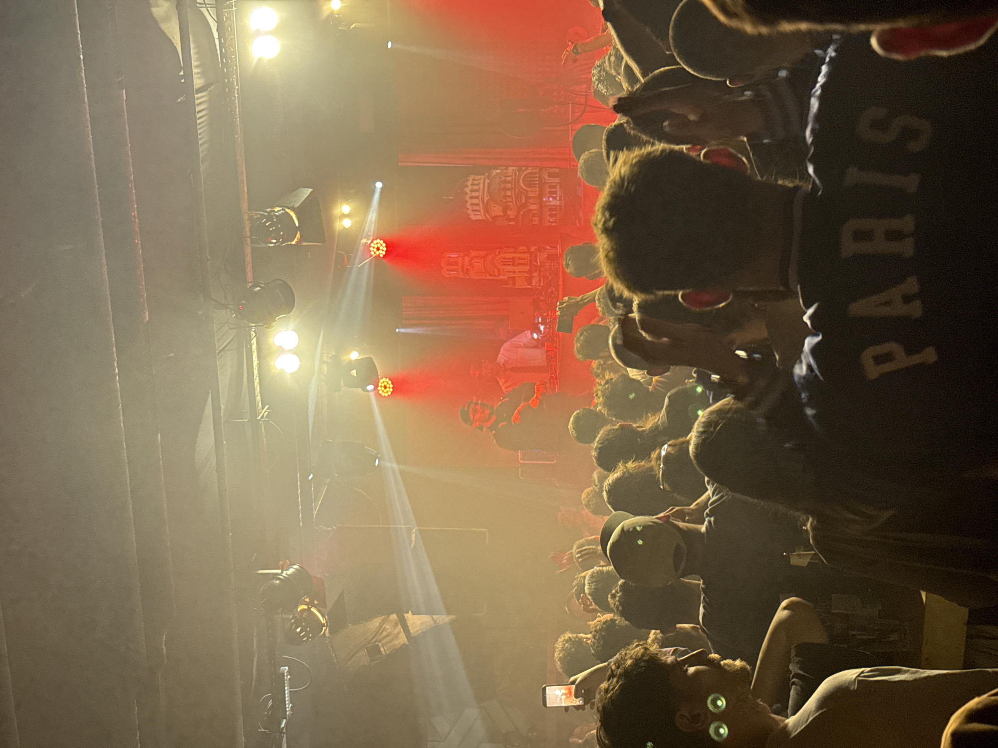 A music concert is taking place in a smoky environment, likely indoors, filled with a crowd of people. The stage features lights and a backdrop structure. A person appears to be performing on the stage. The crowd is facing the stage. A person in the foreground is lying down. The person's shirt says "PARIS". The location is unknown, and there is no verifiable link to Sofia, Bulgaria.