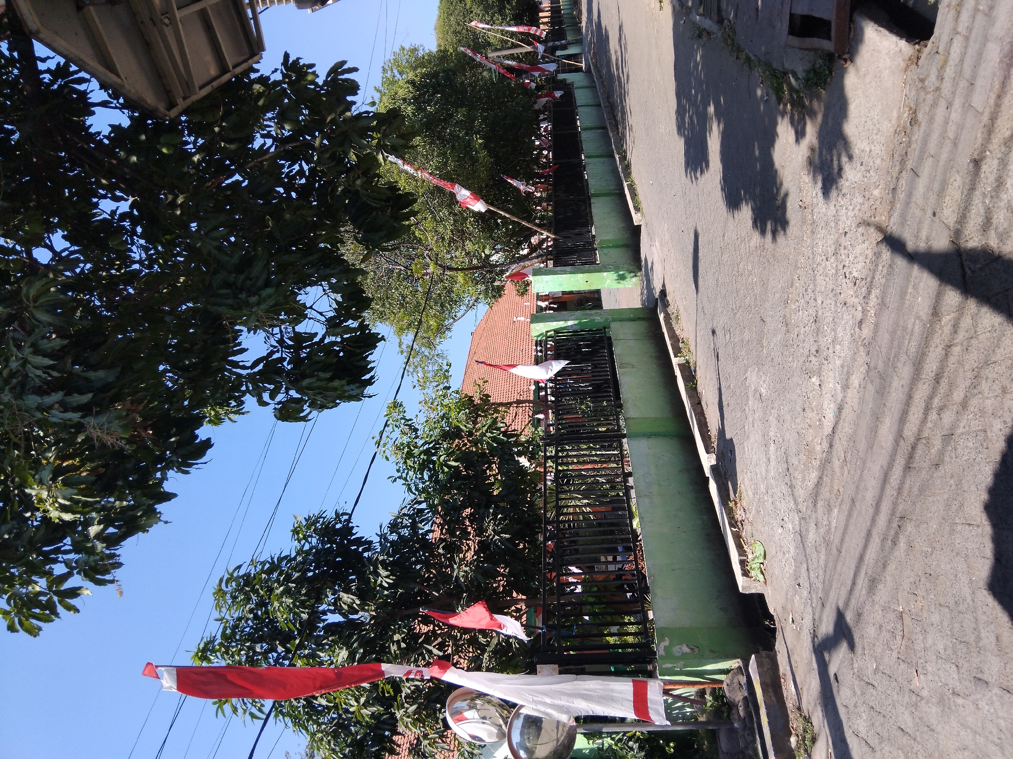 This is a photograph taken outdoors in Berbek, Indonesia, on a clear, sunny day. The scene depicts a street lined with a low, green-painted wall and a black metal fence. Red and white Indonesian flags are prominently displayed along the fence and on poles, suggesting a celebration or national holiday. Trees with lush green foliage are visible behind the fence and overhead, casting shadows on the paved street. The street itself is made of interlocking stone pavers in the foreground and asphalt further down. A building with a red-tiled roof is visible behind the fence, and the edge of a roof or awning is visible in the upper right corner of the frame. There are no people visible in the image, and the focus is on the streetscape and the patriotic decorations.
