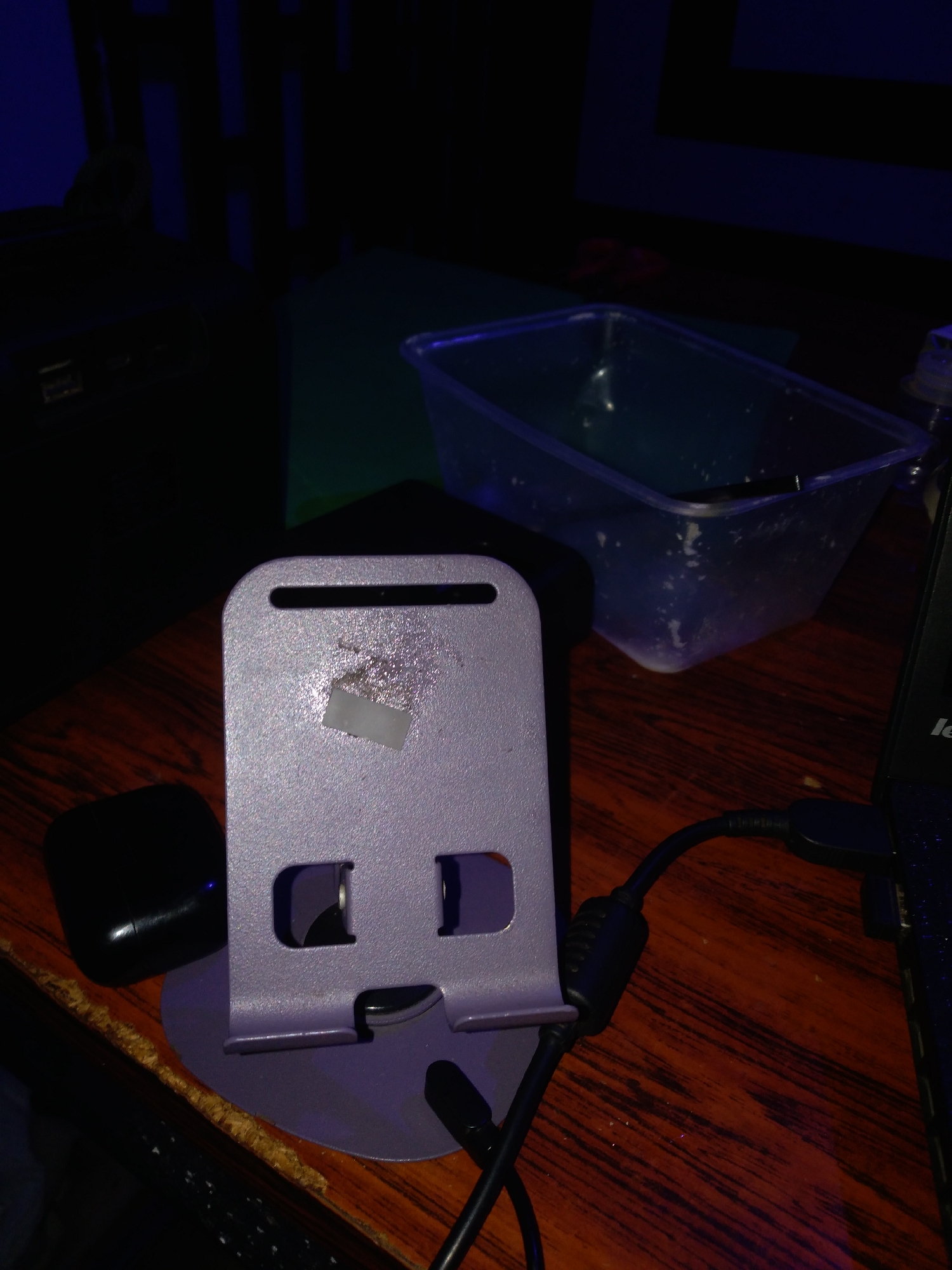 The scene depicts an indoor environment, likely a desk or workstation, under low, predominantly blue and purple ambient lighting, located in Jos, Nigeria. In the foreground, a silver-grey or light purple mobile phone stand with a round base is centrally positioned. A small, rectangular piece of white adhesive residue or tape adheres to its upper surface. To the immediate left of the stand, a black, oval-shaped wireless earbud case with a faint blue indicator light is present. A black cable, equipped with a ferrite bead, exits the base of the phone stand and connects to the right, leading towards a partially visible black Lenovo laptop located on the right side of the frame, its keyboard displaying illuminated keys. Behind the phone stand and partially obscured by it, a translucent plastic container filled with a liquid, likely water, and some submerged dark objects is visible; the container shows internal condensation or residue. All objects rest on a dark reddish-brown wooden surface with a distinct grain, and the left edge of this surface exhibits some wear or chipping. The background is largely dark, with hints of blue illumination highlighting abstract vertical or horizontal dark patterns, potentially on a wall or shelving unit.