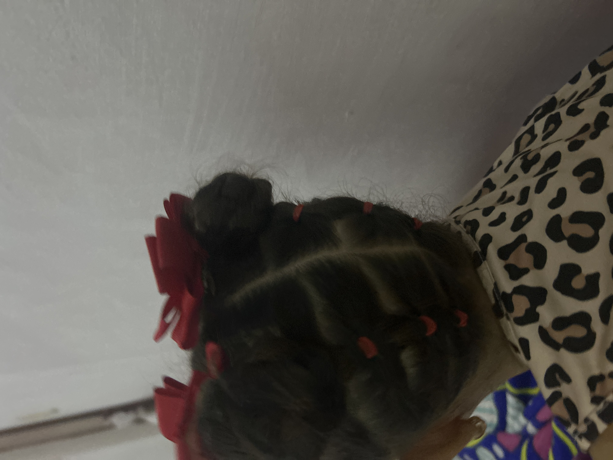 The image shows the back of a child's head with dark hair styled in small buns or sections separated by parts. Small red hair ties or bands secure the sections. A large red bow is attached to one side. The child is wearing an animal-print patterned top. The background is a white wall and part of what appears to be a table. The location is indeterminate, but the request specified San Juan de Los Morros, Venezuela.