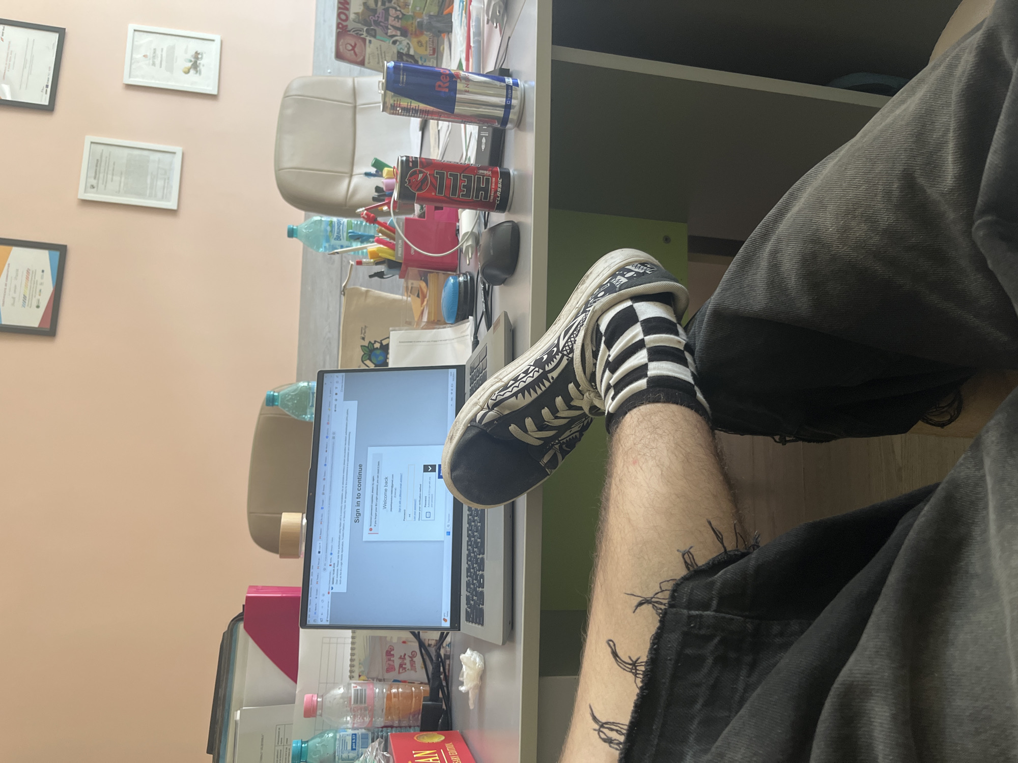 The image depicts a person's legs and feet resting on a desk in what appears to be an office environment. The laptop screen displays a login page. On the desk, there are two energy drink cans, one red labeled "HELL CLASSIC" and another blue and silver labeled "Red Bull ENERGY DRINK". A computer mouse is also visible. In the background, an office chair is partially visible, and framed certificates or awards are mounted on a peach-colored wall. The floor appears to be wooden. There is no indication of the specific location within Vratsa, Bulgaria.